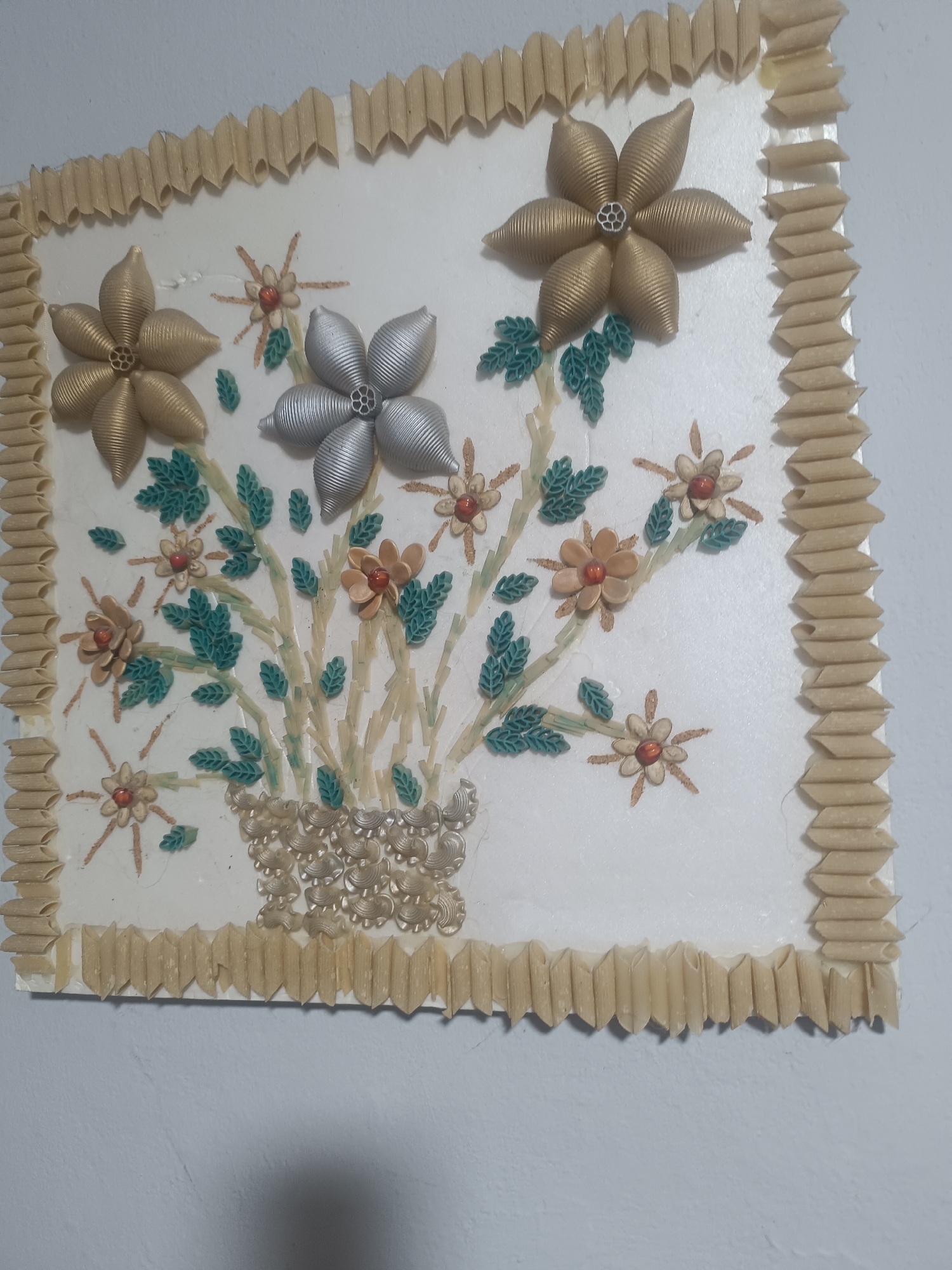 The image displays a wall-mounted art piece crafted from pasta. The central design features a bouquet of flowers, with larger, metallic-painted shell pasta flowers in gold and silver hues, and smaller, tan-colored shell pasta flowers with red bead centers. The stems and leaves are constructed from green and tan-colored pasta pieces. The entire artwork is framed with penne pasta arranged in a zig-zag pattern. The background is a plain white surface, and the artwork is mounted on a wall, possibly in a home or public space within the village of Бәйдібек би ауылы, Kazakhstan.
