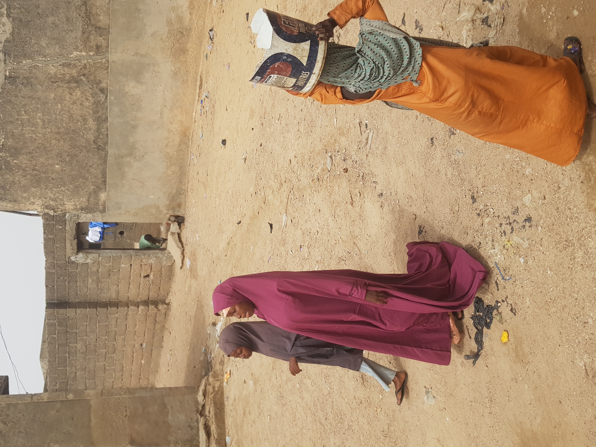 The image depicts a street scene in Dutsinma A, Nigeria. Two women in traditional Islamic dress, one in a dark rose robe and the other in a grey robe and jeans, walk together on a sandy surface. A third woman, in an orange robe, walks in the opposite direction with a printed container on her head. Debris and trash are scattered on the ground. A partially-constructed brick building is on the left, with a figure visible in a window opening along with some hanging clothing.