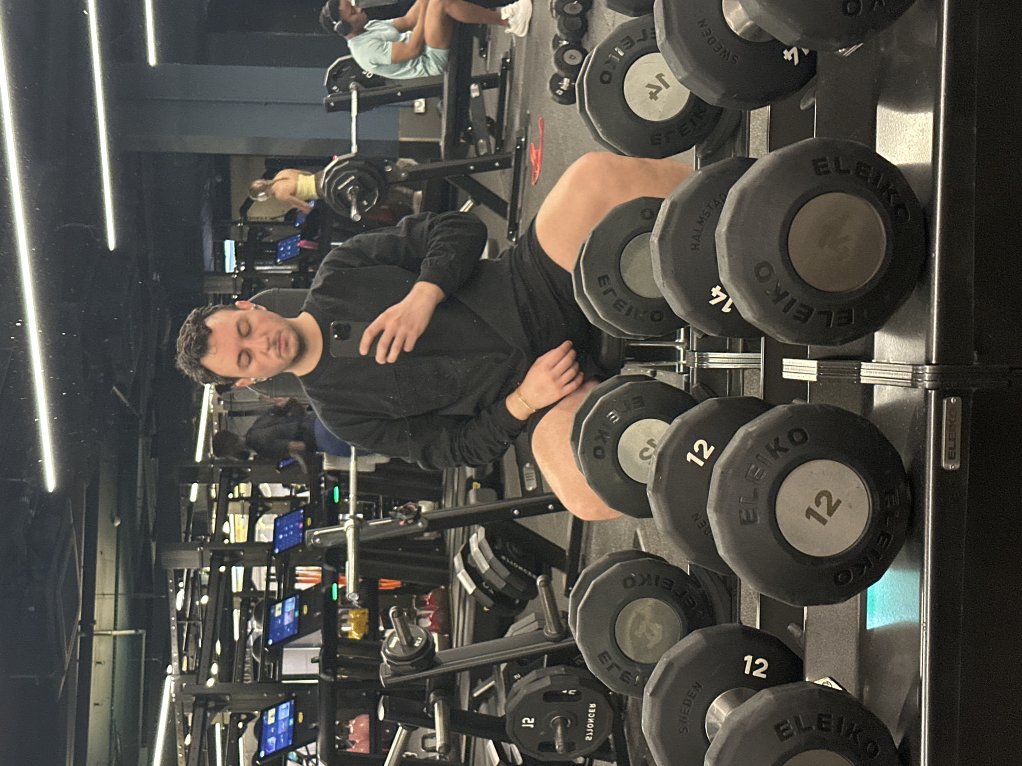 Here is a detailed description of the image:
**Scene:** The image appears to be taken in a gym, possibly in London, United Kingdom, given the presence of "Eleiko" branded weights (a company originating in Sweden, but with a strong presence in gyms worldwide), and the overall look of modern gym equipment often found in London. The image is likely a reflection in a mirror, as indicated by the inverted perspective and the person holding a phone towards the reflection.
**People:**
* One man is clearly visible in the mirror's reflection. He is holding a smartphone, presumably taking the picture. He wears a black long-sleeved shirt and dark shorts.
* Other individuals are vaguely visible in the background, engaged in gym activities.
**Objects:**
* The foreground is dominated by a dumbbell rack holding multiple sets of Eleiko dumbbells of varying weights (e.g., "12"). The brand "Eleiko" and the weight "12" are visible on several dumbbells.
* A smartphone is held by the man in the reflection.
* Various gym machines and equipment are visible in the background.
**Actions and Interactions:**
* The main action is the man taking a photo/video in the mirror reflection.
* Background activity includes people using gym equipment.
**Environment:**
* The environment is a well-lit, modern gym with a variety of exercise machines and equipment. The lighting appears to be artificial, possibly fluorescent or LED.
**Location Details (London, UK - assumed):**
Given the likely use of Eleiko weights and the general appearance of the gym equipment, combined with the assumption of this being a photograph posted by a person living in London, it is probable that the image was taken in a high-end gym facility located in London, UK. However, this cannot be definitively confirmed without further information.
