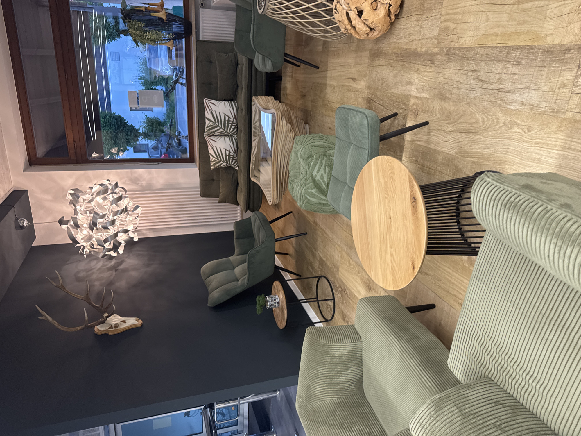 The image shows a modern and stylish seating area in what appears to be a commercial space, possibly a café or lounge, located in Höhenkirchen-Siegertsbrunn, Germany. The room is decorated with a natural and organic aesthetic, featuring light-colored wooden flooring and a dark gray accent wall.
Prominently displayed on the dark wall is a mounted set of deer antlers, suggesting a rustic or nature-inspired theme. Above the antlers, a striking contemporary chandelier hangs, casting a soft light and adding a touch of modern elegance.
The seating arrangement consists of several comfortable-looking chairs and sofas upholstered in a textured, light green fabric. One group of seating is arranged around a round wooden coffee table with a minimalist black metal base. Nearby, a smaller circular side table holds a small potted plant in a decorative metal holder. Another distinctive feature is a uniquely designed coffee table with a layered, curved wooden base and a glass top, reflecting the surrounding environment.
In the background, a large window offers a view of the outdoors, showing trees and some buildings, indicating it is likely daytime. The reflection in the window suggests the exterior is a street or residential area. The overall atmosphere conveyed is one of relaxation and sophisticated comfort. There is no visible text in the image.