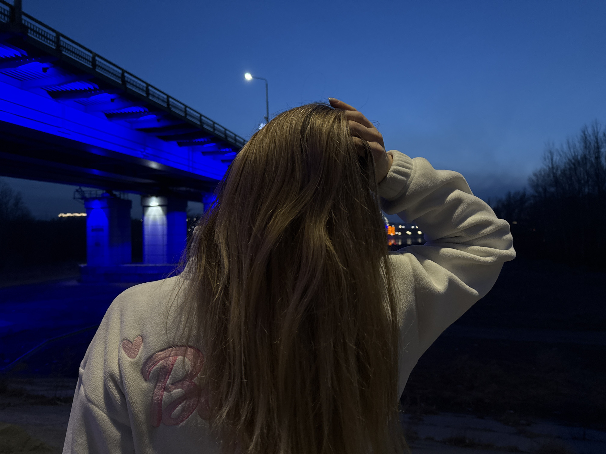 The image captures a person from behind, standing outdoors at night. The individual has long, light brown hair and is wearing a white, textured sweatshirt. On the back of the sweatshirt, there is a pink embroidered logo featuring the word "Barbie" in a stylized font, with a small pink heart positioned above it. One of the person's hands is raised and placed on the back of their head or in their hair.
In the background, a large bridge is visible, illuminated distinctly by bright blue lights underneath its structure and on its support pillars. The sky above is a deep blue, characteristic of nightfall or early evening. A single, bright street light on a tall pole stands out against the dark sky to the left of the person's head. To the right, distant trees or bushes are silhouetted against the horizon, and faint, out-of-focus lights suggest an urban or populated area further away. The foreground and ground directly beneath the bridge are dark and unlit. The scene depicts a calm, nighttime setting, possibly in an urban or suburban environment, with the blue-lit bridge as a striking architectural feature.