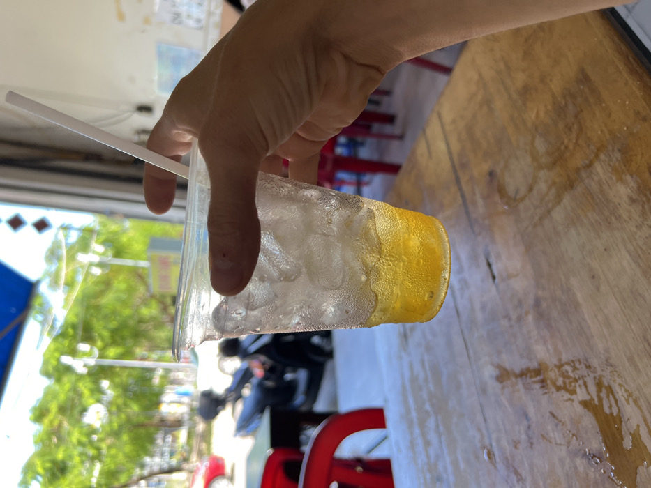 A hand holds a clear plastic cup filled with ice and a yellow liquid, condensation forming on the outside. A white straw sticks out from the top. The background shows a blurry street scene with trees and motorcycles. A wooden table is partially visible on the right. Red plastic chairs are interspersed throughout the background.
