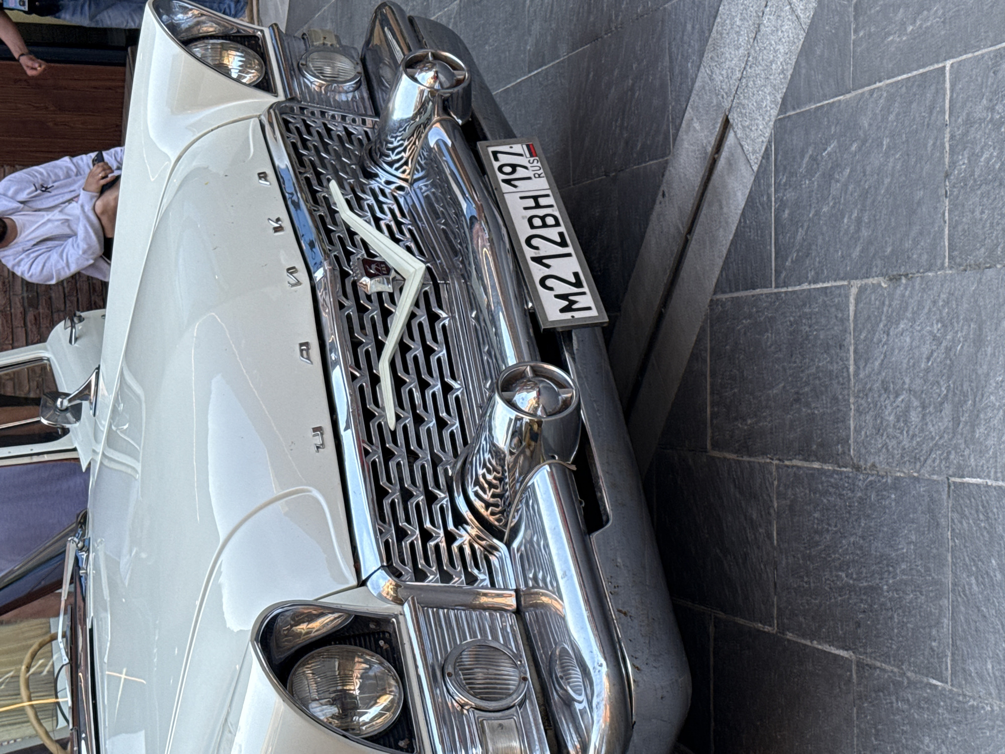 The image shows the front of a white vintage car parked on a gray tiled surface in an urban environment. The chrome grille and bumper are prominent, reflecting the surrounding light. A license plate reading "M212BH 197 RUS" is attached to the front. A person wearing a white jacket is partially visible in the background.