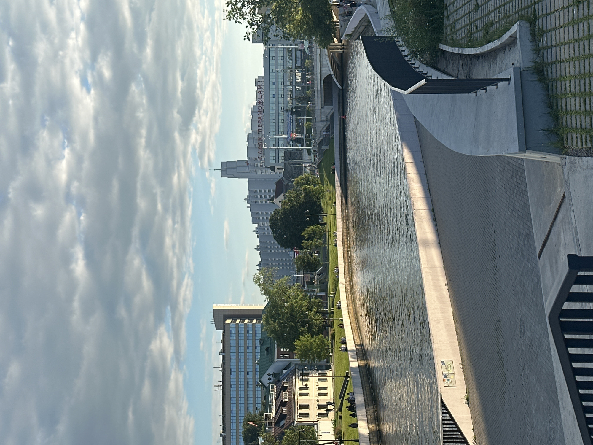 This is an eye-level, medium-shot photograph taken outdoors in the city of Minsk, Belarus, depicting a riverside embankment with a modern cityscape in the background. The scene captures a sunlit day with a partly cloudy sky.
In the foreground, a paved walkway with a textured surface, likely cobblestones, curves along the edge of the river. A low, grey concrete wall separates the walkway from the water, and a black metal railing runs along the outer edge of the walkway. The river itself is a wide expanse of water reflecting the light and sky, with subtle ripples on its surface.
Along the far bank of the river, there is a grassy embankment dotted with people. These individuals are casually dressed and are seen sitting, standing, and walking, enjoying the riverside setting. Behind the embankment, a mix of buildings is visible. To the left, a lower-rise building with white walls and a red-tiled roof stands out, with trees partially obscuring it. Further back, a larger, modern building with a blue-tinted glass facade dominates the skyline, and another taller, rectangular building with a beige exterior is positioned behind it. In the center of the background, a prominent modern building features red Cyrillic lettering that reads "СЛАВА ПЕРЕМОЖЦАМ!" (Glory to the Winners!). A bridge arches over the river in the mid-ground, connecting the two sides of the city. The overall impression is one of an urban environment integrating green spaces and water features with contemporary architecture.