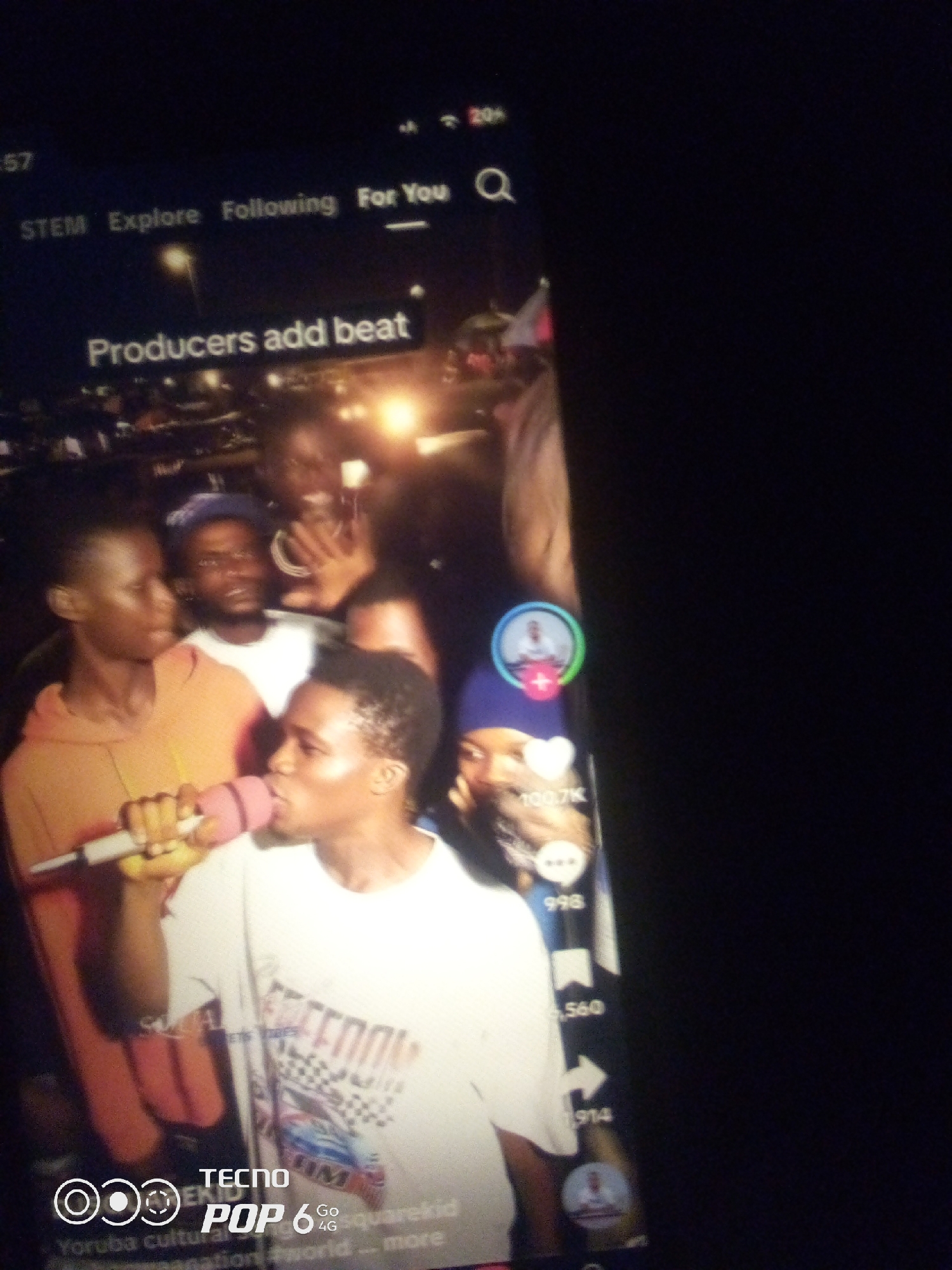 The image shows a close-up of a smartphone screen displaying a social media video. The video appears to be a performance, with a young man in the foreground holding a pink microphone to his lips and singing. He is wearing a white t-shirt with a graphic design. To his left, another young man in an orange hoodie is visible. In the background, other individuals are present, and the setting appears to be an outdoor event at night, possibly in Howayi, Nigeria, suggested by the presence of "TECNO POP 6" branding, a mobile phone brand popular in Africa. The social media interface shows a title "Producers add beat" and engagement metrics such as likes and shares. The overall scene is dark, with artificial lighting illuminating the subjects.