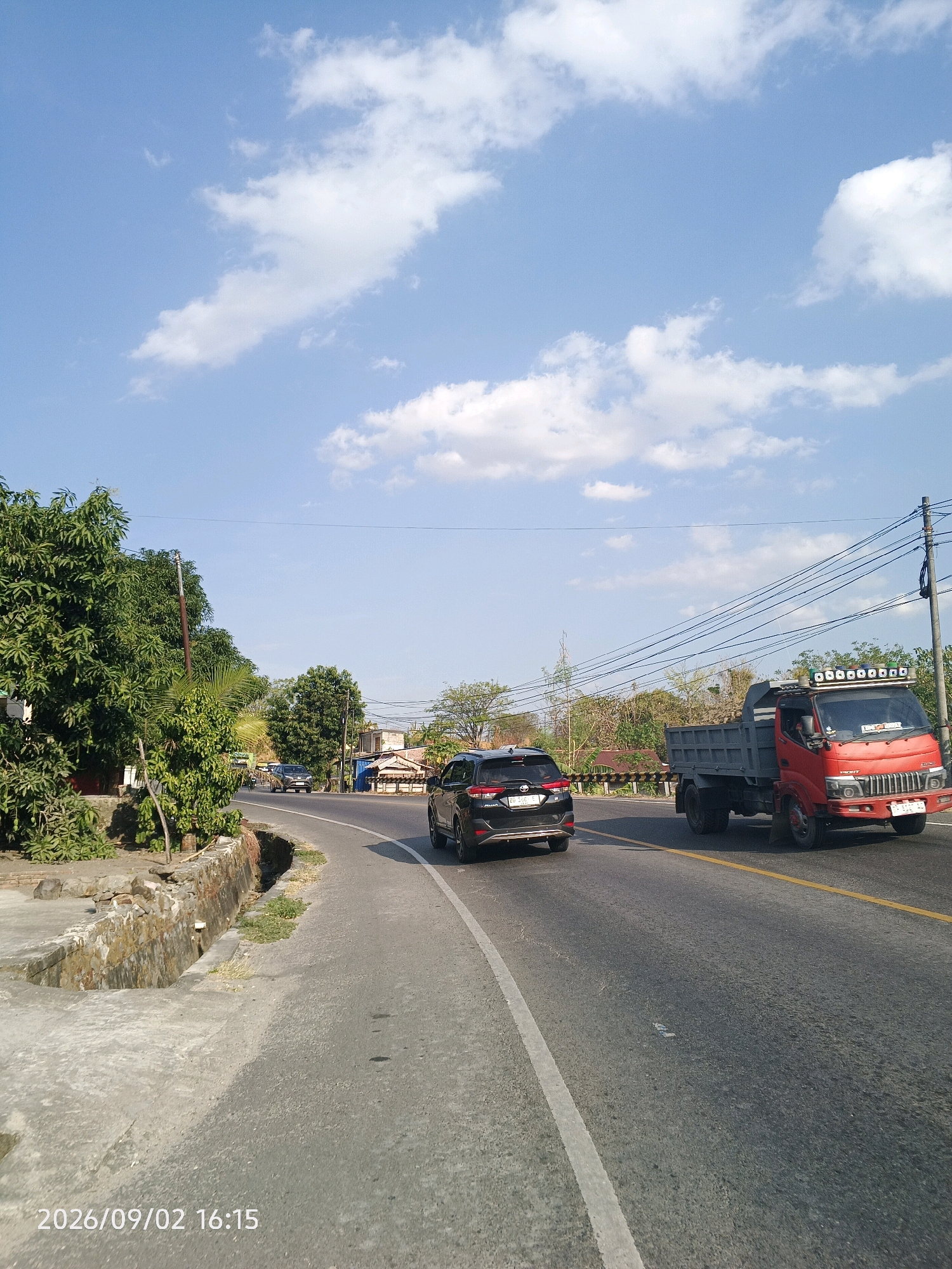 This is a street-level, landscape-oriented photograph taken in Pinrang, Indonesia, on a clear day with scattered white clouds. The road appears to be a two-lane highway, with a yellow line dividing the lanes. A black SUV is traveling in the right lane, moving away from the camera, while a red dump truck is in the left lane, also moving away. The road curves gently to the right in the foreground and continues straight into the distance.
To the left of the road, there is a concrete embankment with some vegetation growing on it. A large tree with green foliage dominates the left side of the frame, with some smaller trees and buildings visible behind it. Power lines stretch across the upper portion of the image, supported by utility poles.
The ground in the foreground is asphalt, with white lane markings. The timestamp "2026/09/02 16:15" is visible in the bottom left corner of the image. The overall scene suggests a typical day on a road in a moderately developed area.