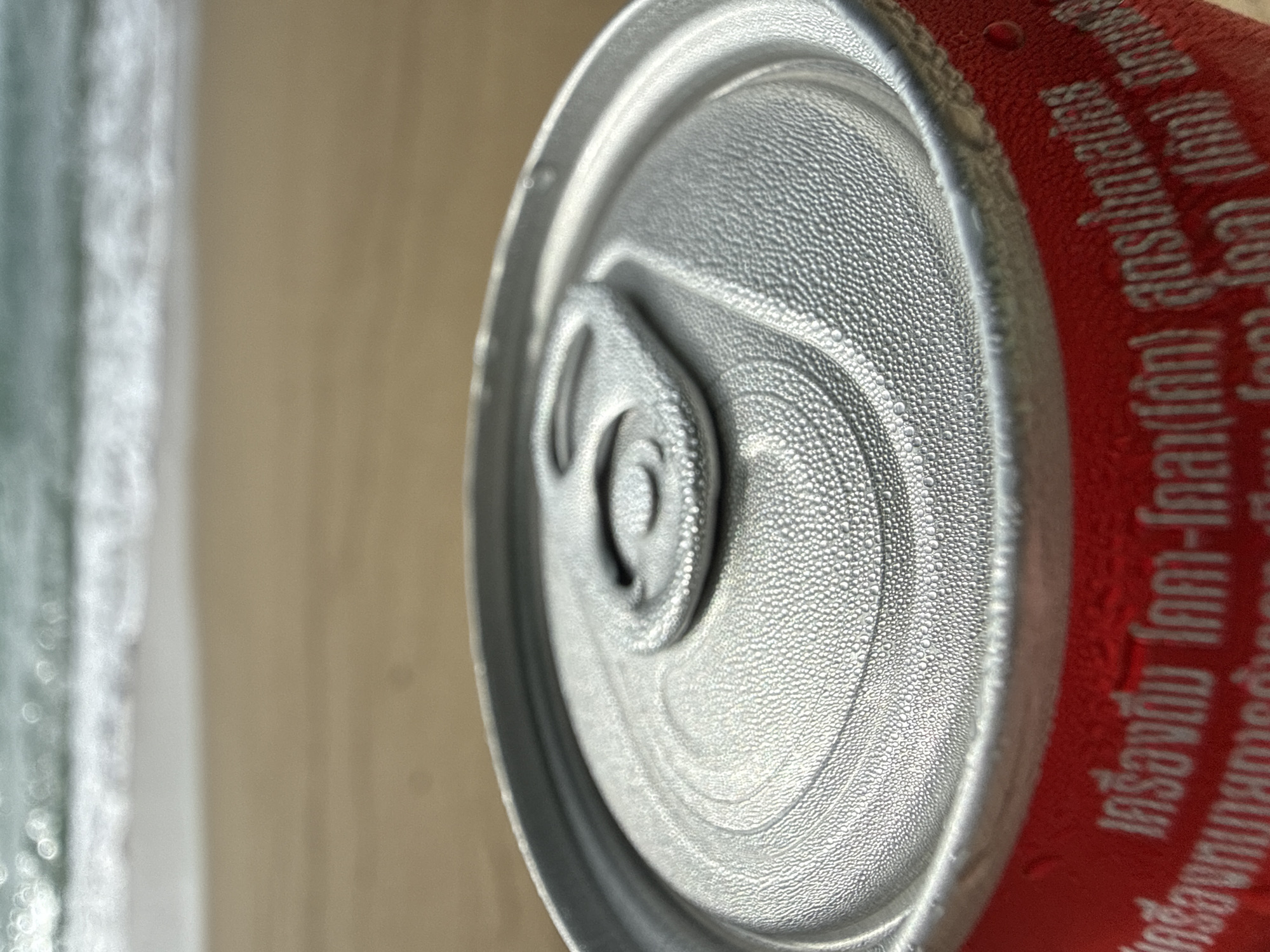A close-up shot shows the condensation-covered top of a red soda can, likely Coca-Cola given the iconic red color and familiar can shape. The can is placed on a sandy beach, with the blurred turquoise ocean and white waves in the background. The text on the can appears to be in Thai, suggesting its location in Thailand. The image captures a moment of leisure, with the beverage offering a refreshment in a tropical setting. The scene suggests a relaxed atmosphere, typical of a beach day in Kamala, Thailand.