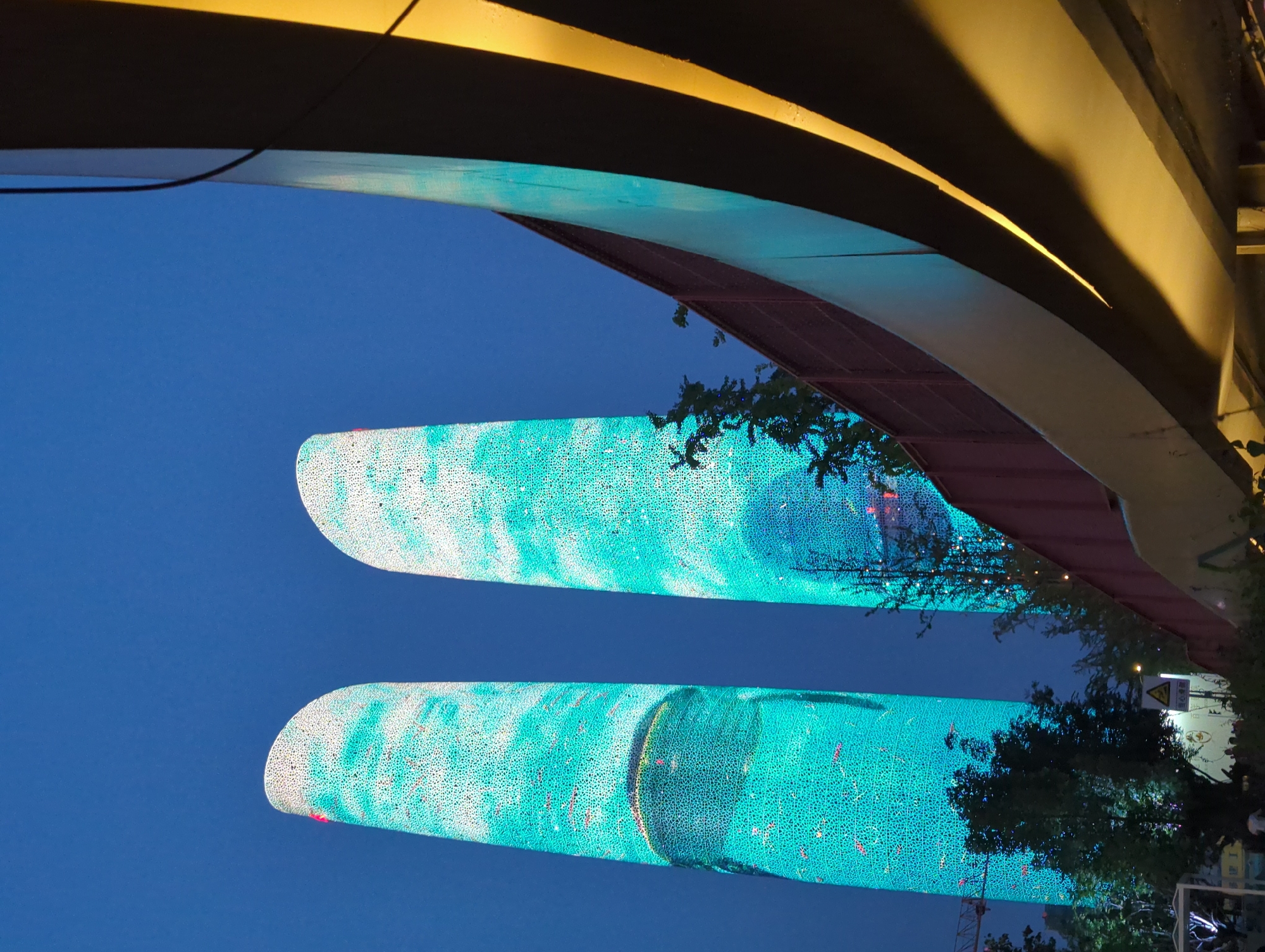 The image depicts the iconic Twin Towers of Chengdu, located in the Wuhou District, China, during twilight. The skyscrapers are illuminated with a vibrant teal and white light display, creating a striking visual against the deep blue sky. A curved, overpass-like structure dominates the foreground, casting dramatic shadows and highlighted with a warm yellow light. Trees and foliage are visible at the base of the towers, adding a touch of nature to the urban landscape. A construction crane can be seen in the distance to the left. The scene appears to be set in a public area, with hints of pedestrian activity at the lower left of the frame, though no individuals are clearly discernible. The overall impression is one of modern architecture and urban beautification.