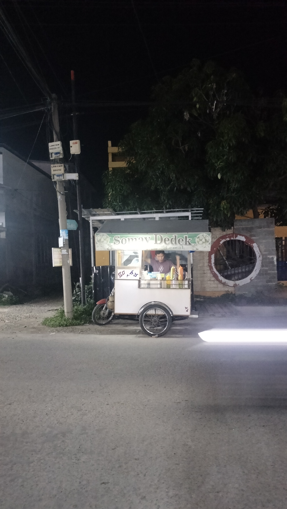 An outdoor, nighttime scene in Lhokseumawe, Indonesia, depicts a street-side food cart selling "Somay Dedek Lemak." The cart, a white three-wheeled vehicle, is illuminated by a bright light emanating from within and also by ambient streetlights. A man, presumed to be the vendor, is visible inside the cart, wearing a purple t-shirt and with his arms raised. He is surrounded by containers of condiments, including yellow and orange sauces. The cart is parked on the side of a paved road, with a small patch of green vegetation to its left. In the background, a dark tree with some hanging fruit is prominent, and beyond that, a textured wall with a circular opening is visible. A utility pole with electrical wires and several boxes attached stands to the left of the cart. The overall impression is a common urban scene at night, highlighting a local food business.