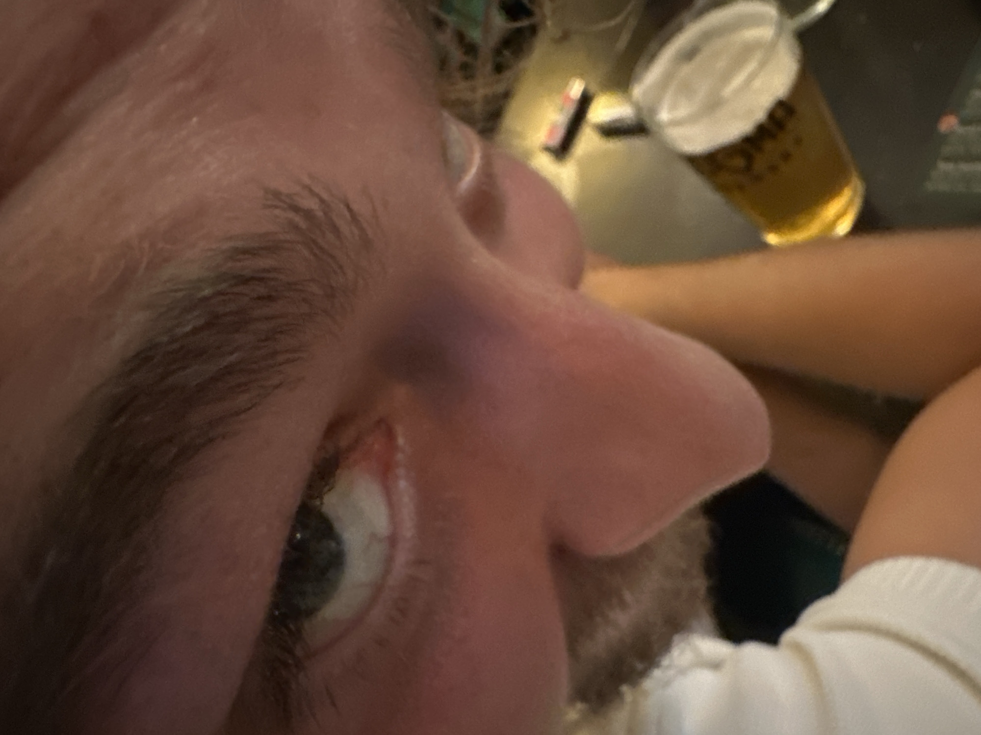 A close-up, low-angle shot captures a person's face and a glass of beer on a table. The person's right eye, with a green iris, is visible. The nose is prominent and illuminated, with some facial hair on the chin. The person is wearing a white garment with a ribbed texture, possibly a shirt or sweater. In the background, a glass filled with a golden beverage, likely beer, sits on a dark surface. A small electronic device with buttons is also partially visible on the table. The setting appears to be indoors, with artificial lighting casting warm tones on the skin. There is no discernible information within the image to indicate a specific location in Belgrade, Serbia.