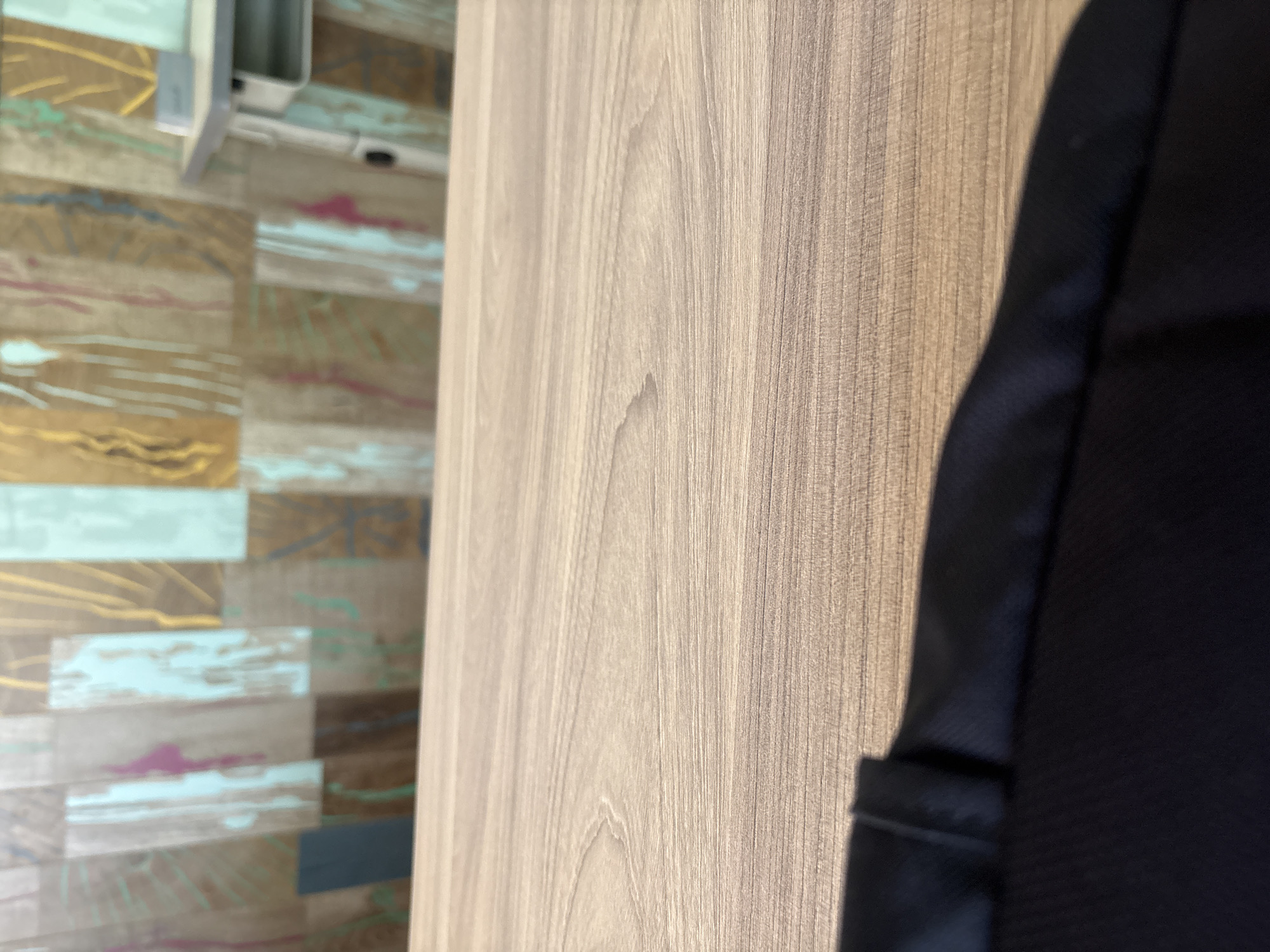 The image displays a close-up, low-angle shot of a wooden surface, likely a table, with a black fabric object partially visible in the foreground. In the background, a wall with a colorful, abstract wood-paneled design is out of focus, featuring vertical planks in shades of brown, teal, and pink. A blurred portion of a desk with a light-colored top and a gray drawer is also visible to the right, suggesting an indoor setting, possibly a classroom or office in Redina, Bulgaria. No people or specific actions are discernible.