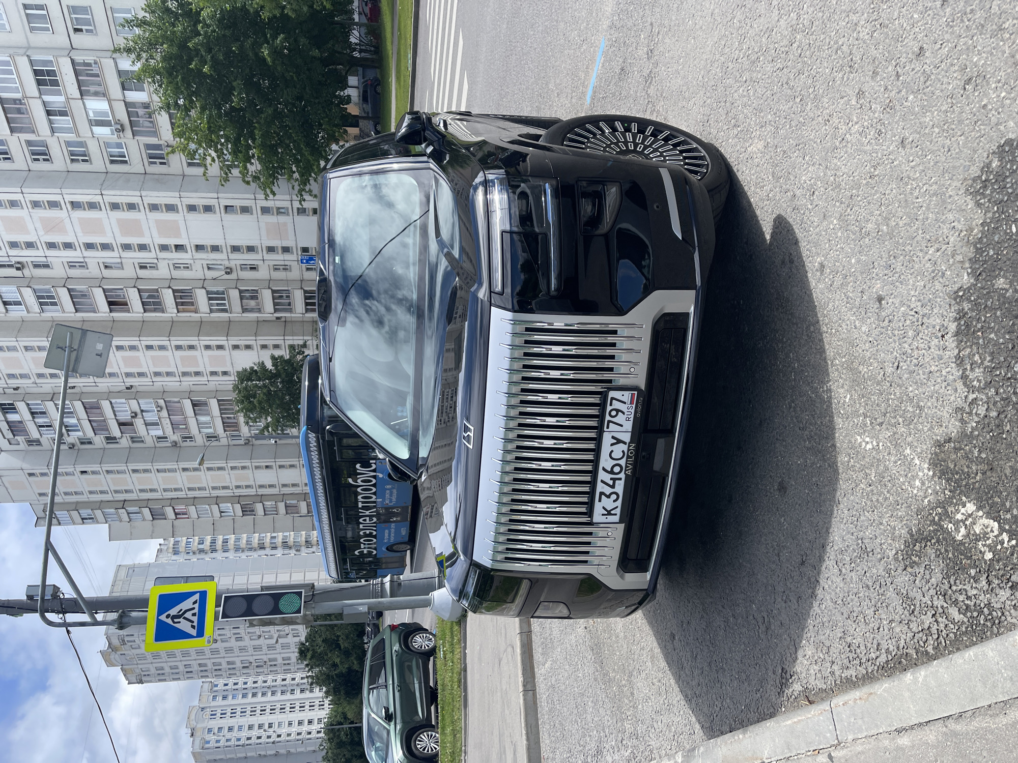 This is a close-up, eye-level, medium shot of a black luxury SUV parked on a street in Moscow, Russia. The car is positioned at an angle, facing slightly towards the viewer's left, with its front grille and headlights prominently displayed. The SUV has a large, vertical chrome grille with thin slats and a silver trim. Its headlights are sleek and modern, with horizontal LED accents. The license plate is visible and reads "K346CY 797 RUS".
In the background, several multi-story apartment buildings with light-colored facades and many windows are visible. A blue public bus with text in Russian ("Это электробус" which translates to "This is an electric bus") is also present in the background, suggesting an urban setting. A pedestrian crossing signal with a green walking person icon is illuminated, and a pedestrian crossing sign is mounted on a traffic light pole. The street is paved with asphalt, and a concrete curb runs along the left side of the image. Green trees and other vehicles are also visible in the distance. The sky is partly cloudy.