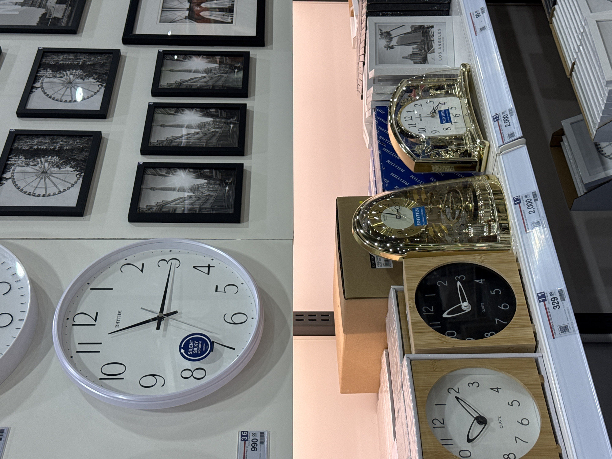 The image depicts a retail display of clocks and framed pictures in a store located in Baan Chaweng Noi, Thailand. A large white wall clock with black numerals and hands, branded "RHYTHM," is prominently displayed. Below this, shelves are stocked with various items. On the top shelf, framed black and white cityscapes and a Ferris wheel are visible. The middle shelf contains cardboard boxes and a gold-colored mantel clock with a glass dome, also branded "RHYTHM." To the right, there are framed pictures with palm trees and city skylines, labeled "LOS ANGELES." The lower shelves display more clocks with wooden frames, one with a white face and the other with a black face, both featuring "QUARTZ" movement. Pricing tags are attached to some of the items, indicating prices in Thai Baht. The overall environment is that of a well-organized retail space.