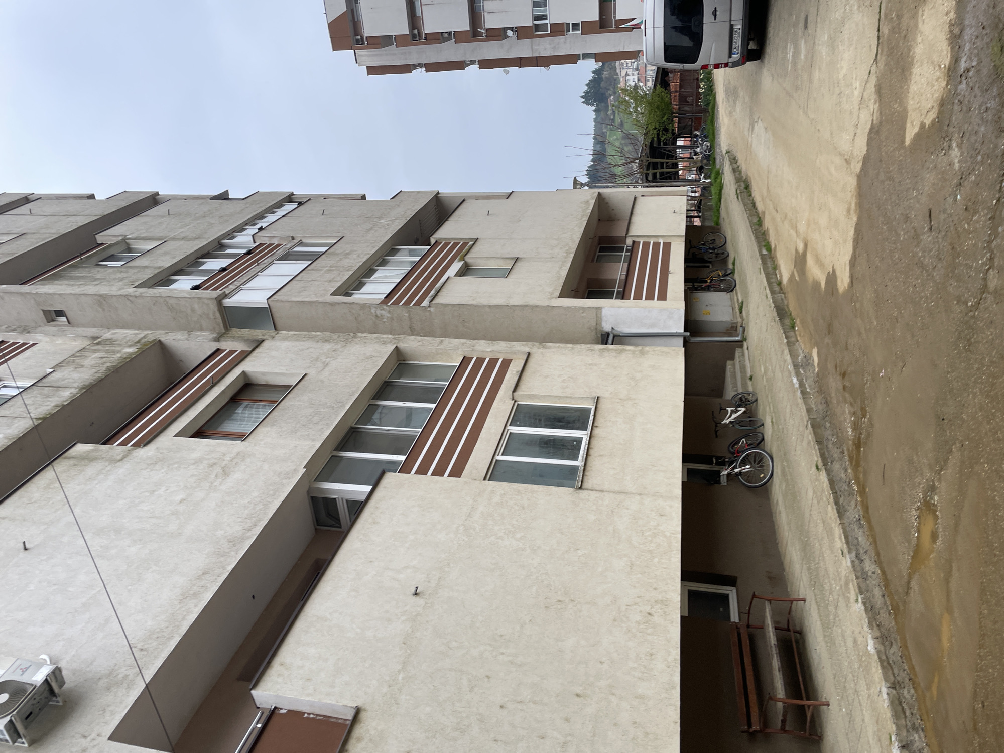 The image depicts an exterior view of a building in Zlatograd, Bulgaria. The building is constructed of concrete with textured surfaces and a beige or off-white color. The architectural style is characterized by protruding, cantilevered sections that create a staggered façade. Windows are rectangular and arranged in horizontal bands across the façade. Brown horizontal stripes are visible between some sets of windows.
Adjacent to the building is a concrete paved alleyway or street, which appears to be wet. Bicycles are parked along the wall of the building, within the alley. In the background, a hill or mountain is visible, with greenery and buildings dotting its slope. Another building can be seen above the main building in the image. A white van is partially visible at the upper right of the image. The sky is overcast. An air conditioning unit is mounted on the exterior of the building.