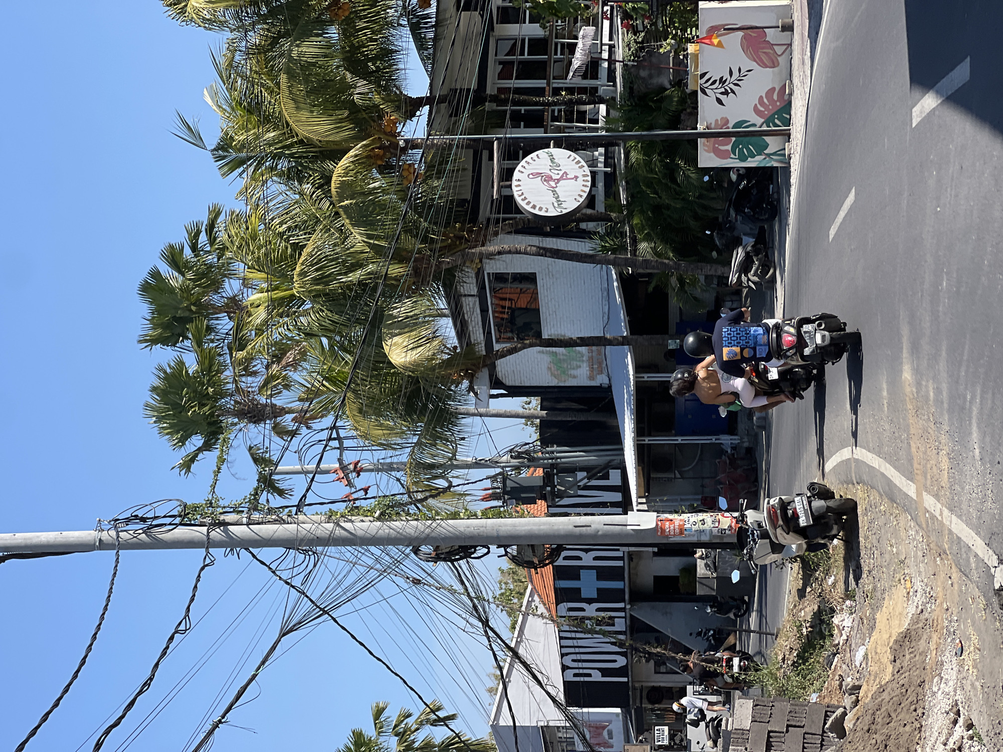 The image captures a street scene in North Kuta, Indonesia, under a clear blue sky. A paved road curves to the right, with a motorbike carrying two passengers, a male and a female, in the foreground, traveling away from the viewer. The female is wearing a light-colored top and the male is wearing a dark blue t-shirt with a graphic print. Both are wearing helmets. To the left of the road, a pile of bricks and dirt is visible, with a motorbike parked nearby. Further down the road, several other motorbikes are parked or in motion, suggesting a busy area.
On the left side of the street, a building with "POWER + RIVE" in large black letters on its facade is prominent. Adjacent to it, another building has a sign that reads "VILLA FOR RENT".
On the right side of the street, a white building with a balcony features a circular sign with a flamingo graphic and text that reads "COWORKING SPACE TROPICAL NOMAD CANGGU - BALI". Palm trees with coconuts are interspersed throughout the scene, their fronds reaching towards the sky. A utility pole with a complex network of black wires is a dominant feature on the left side of the image, with a transformer box attached to it. A white wall adorned with a tropical leaf and flamingo mural is visible on the far right. The overall impression is a lively, tropical urban environment.
