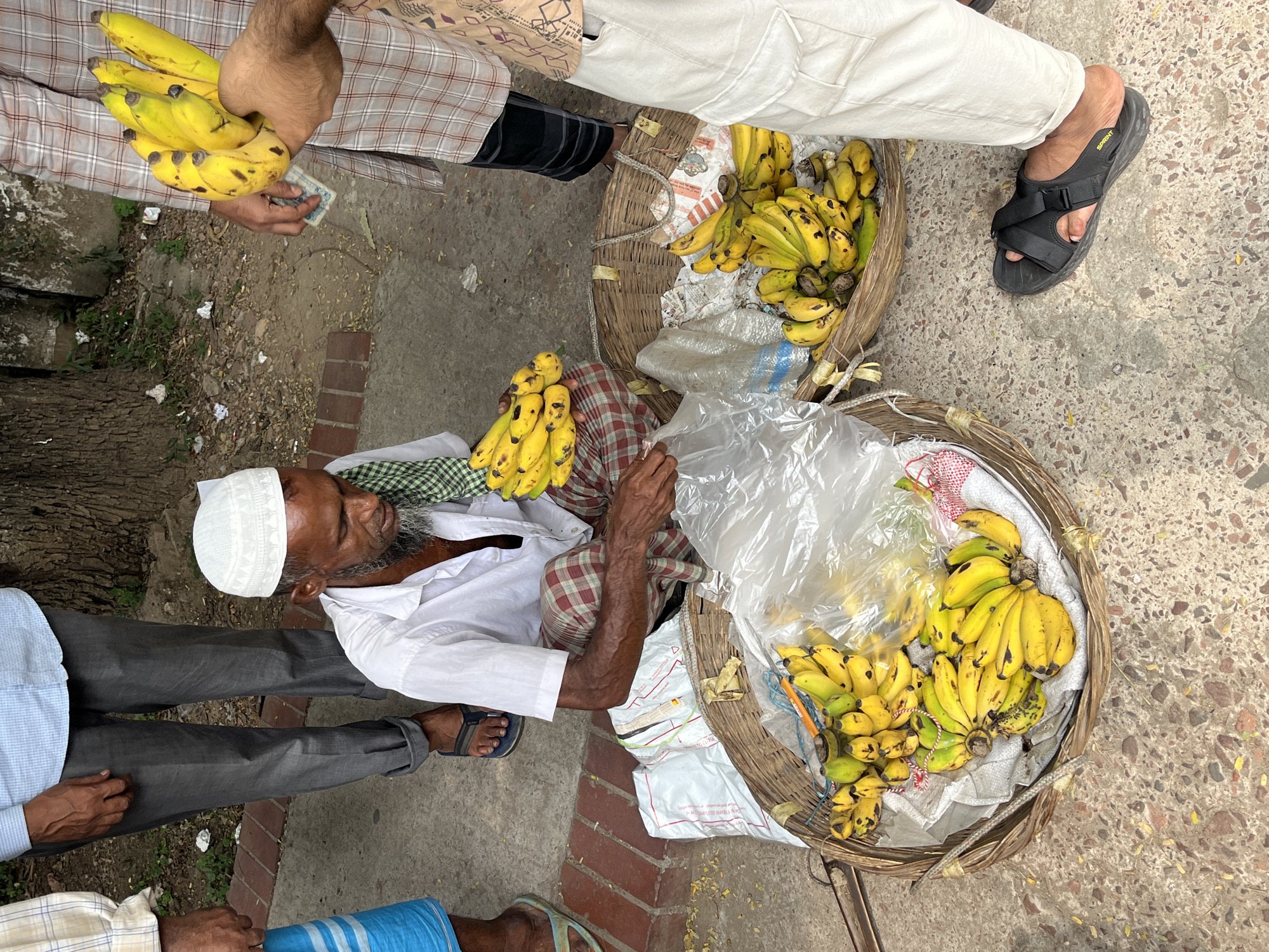 An aerial view shows a banana vendor sitting on the ground in Dhaka, Bangladesh. He is an elderly man wearing a white shirt and a traditional Muslim cap, surrounded by baskets filled with yellow bananas. Customers stand around him, one of whom holds a bunch of bananas in his hand, seemingly making a purchase. The setting appears to be a roadside market or street vending area, with the ground made of concrete slabs and brick.