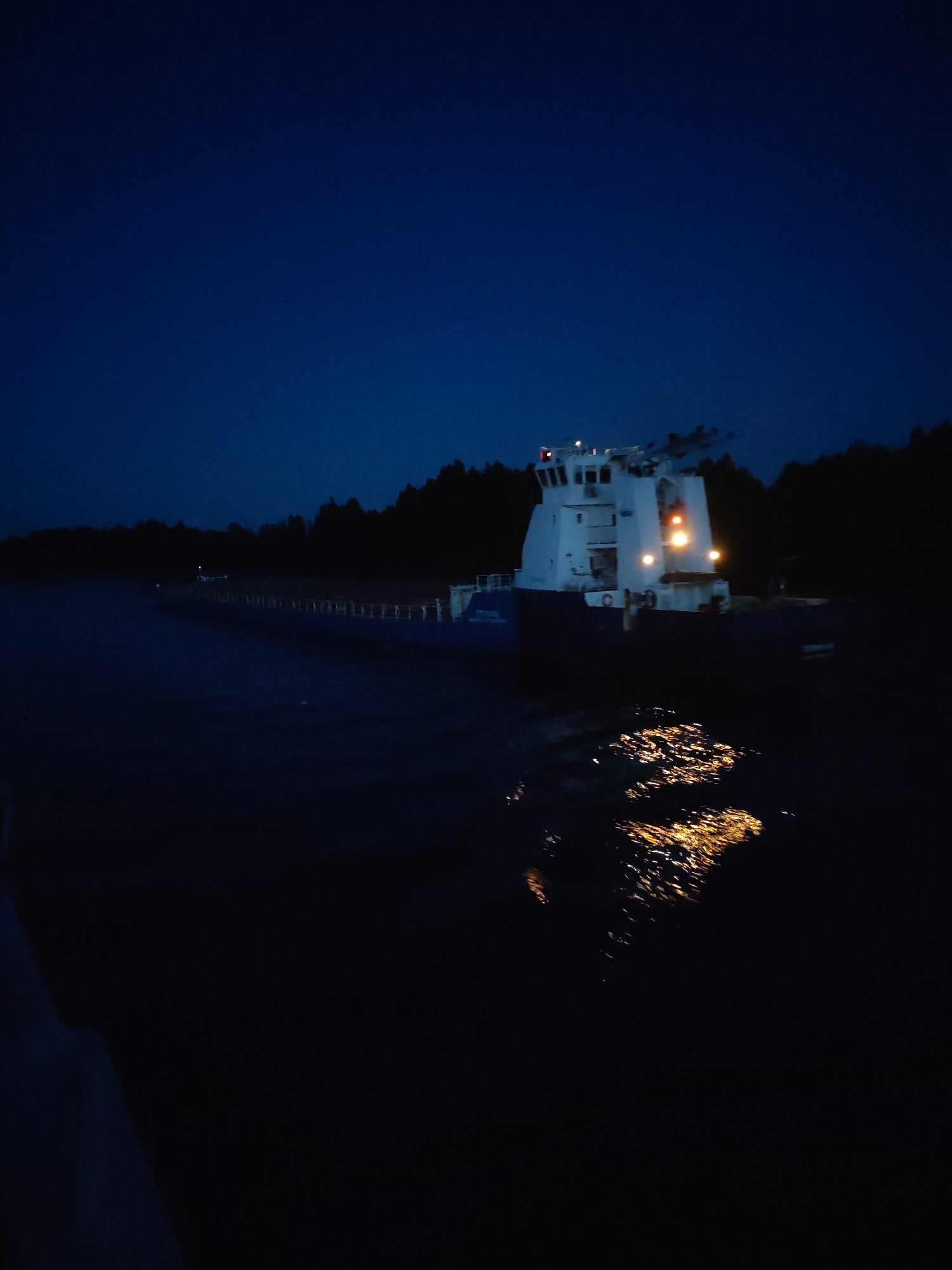 The image depicts a vessel on the dark waters of the Vytegra River in Vytegra, Russia, during twilight or nighttime. The sky is a deep, clear blue. A white superstructure with illuminated windows and lights is visible on the vessel, which is positioned on the right side of the frame. The water's surface reflects the lights from the vessel, creating a shimmering effect. A dark, tree-lined shore is visible in the background. The overall atmosphere is quiet and serene.