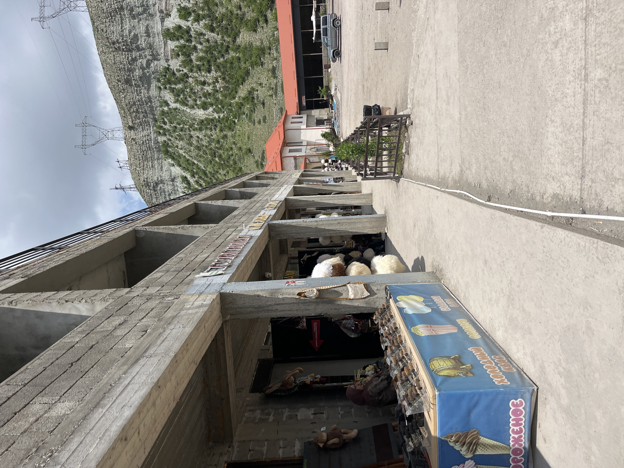The image depicts a street scene in Chirkey, Russia. A multi-story, unfinished concrete building dominates the left side of the frame, its raw construction visible with open window spaces. At ground level, a shop is open, displaying various items including stuffed animals and souvenirs. An ice cream advertisement with Russian text is positioned at the edge of the shop's storefront. Higher up on the building's floors, there are displays of sheepskin hats or similar items.
In the background, there is a mountain covered with vegetation and power lines crossing the sky. Further back, there is a paved area with a building featuring a red roof and at least one vehicle.
The ground is concrete. A white cable runs along the ground.