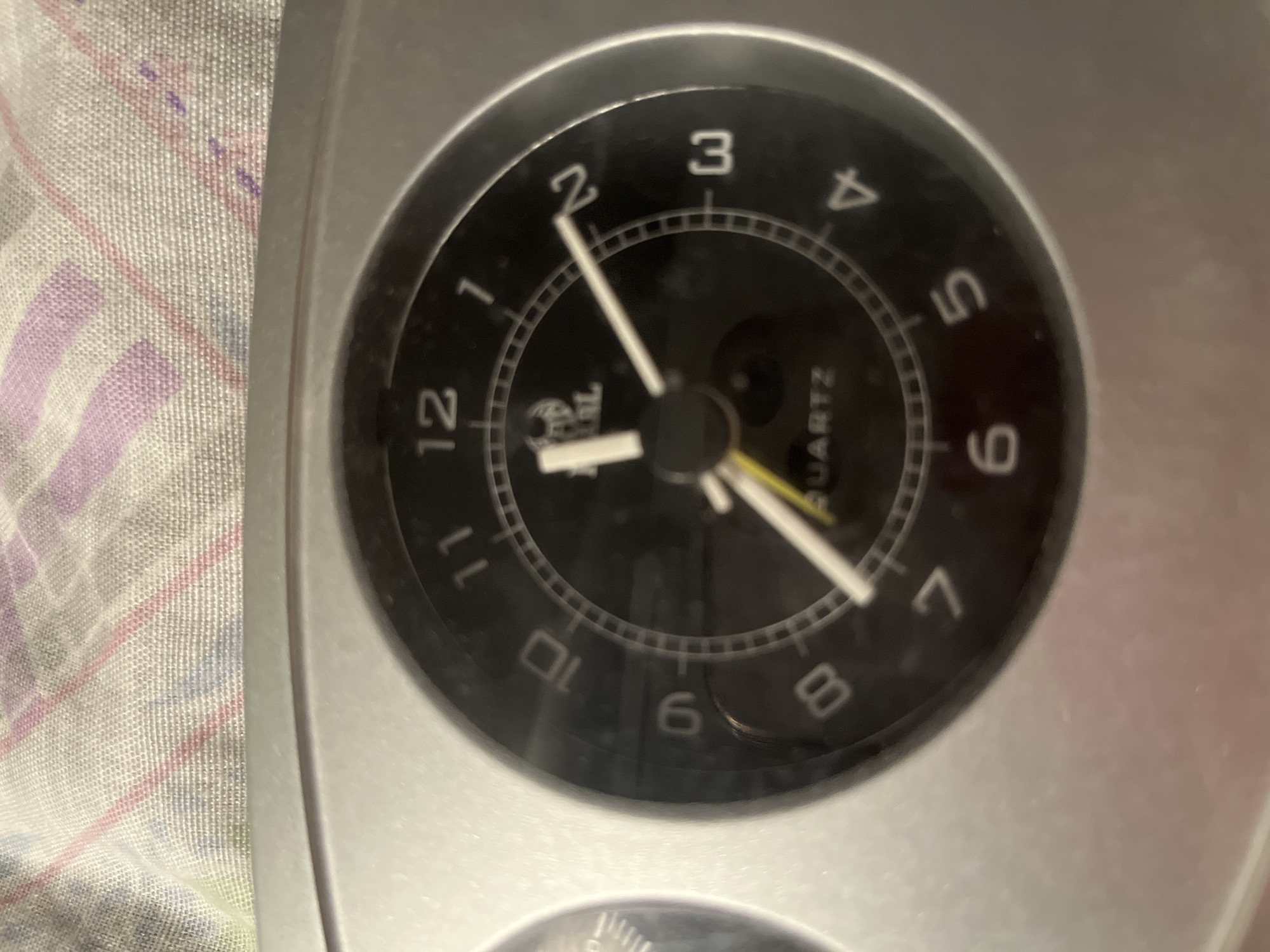 The image shows a close-up of a black analog clock face with white numbers and hands. The clock is set against a textured, fabric-like background with pink and purple stripes. The numbers on the clock face range from 1 to 12. The hour hand is short and white, pointing slightly past the 1. The minute hand is longer and white, pointing to the 2. A thin yellow second hand is also visible, near the 1. The word "QUARTZ" is printed in white text on the lower half of the clock face. The clock appears to be part of a larger object, possibly an alarm clock or a dashboard, as a sliver of a second dial is visible on the left side of the image. The overall lighting suggests the photograph was taken indoors.