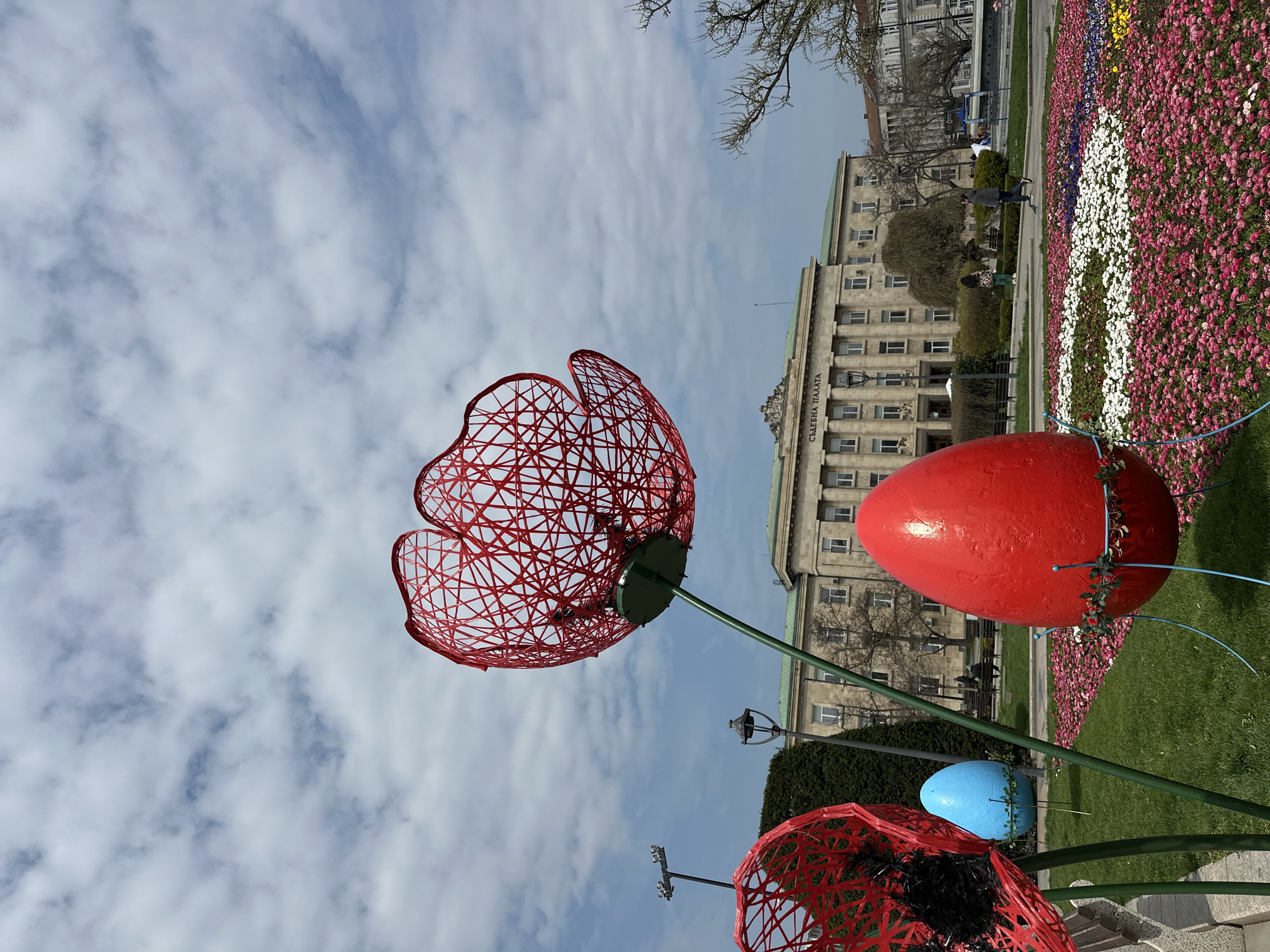 A low-angle, outdoor shot presents a scene in Ruse, Bulgaria. The foreground is dominated by large, artificial flower and egg decorations, part of an Easter installation, suggesting a celebration or event. The flowers are constructed of red wire mesh, creating a skeletal outline, while the eggs are painted in vibrant colors, including red and blue.
Behind the decorations, a well-manicured green lawn extends towards a flower bed arranged in a pattern. Beyond the flower bed, a stone building is visible, identified by the inscription "СЪДЕБНА ПАЛАТА", indicating it is a courthouse or similar government building. Pedestrians can be seen on the sidewalk in front of the building, suggesting daily activity in the area.
The sky overhead is filled with white and gray clouds. The overall scene is a public park or square in Ruse, adorned with Easter decorations.