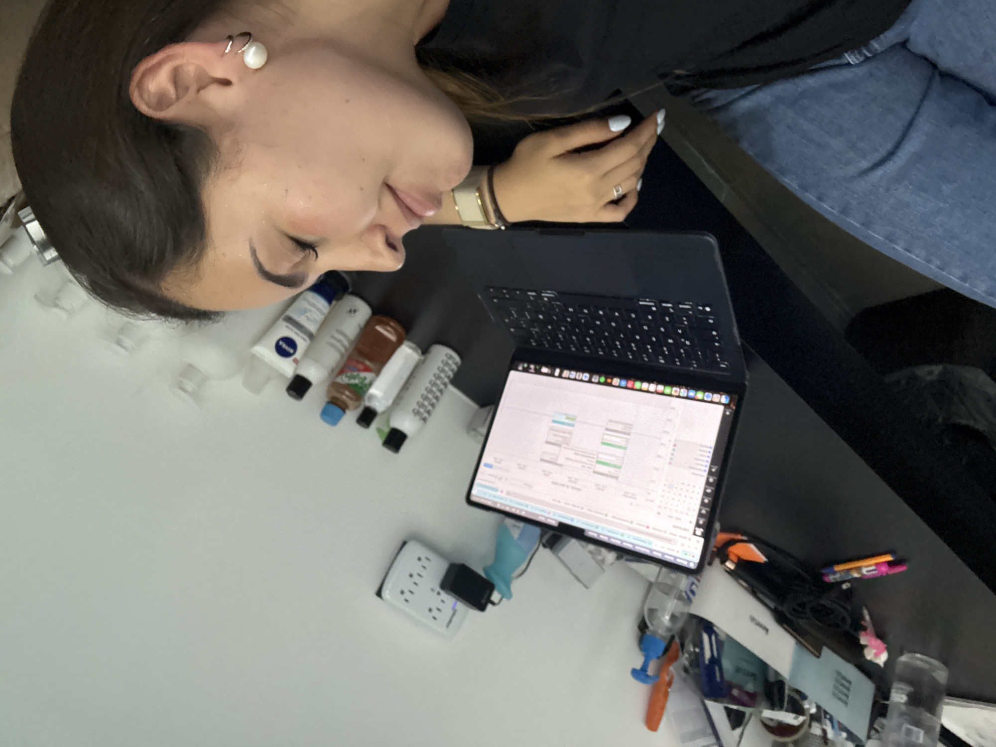 A woman is reclining, her head resting near an assortment of lotion tubes. She is wearing a wristwatch and a ring on her left hand. Her right hand is positioned near an open laptop with a keyboard. The screen displays a spreadsheet-like document. A surge protector with a black adapter plugged in is visible near the lotions. Other objects clutter the desk, including pens, bottles, and cables. The woman is wearing blue jeans and a black top. There's not sufficient information to provide details on the location in San Bernardino Tlaxcalancingo, Mexico.