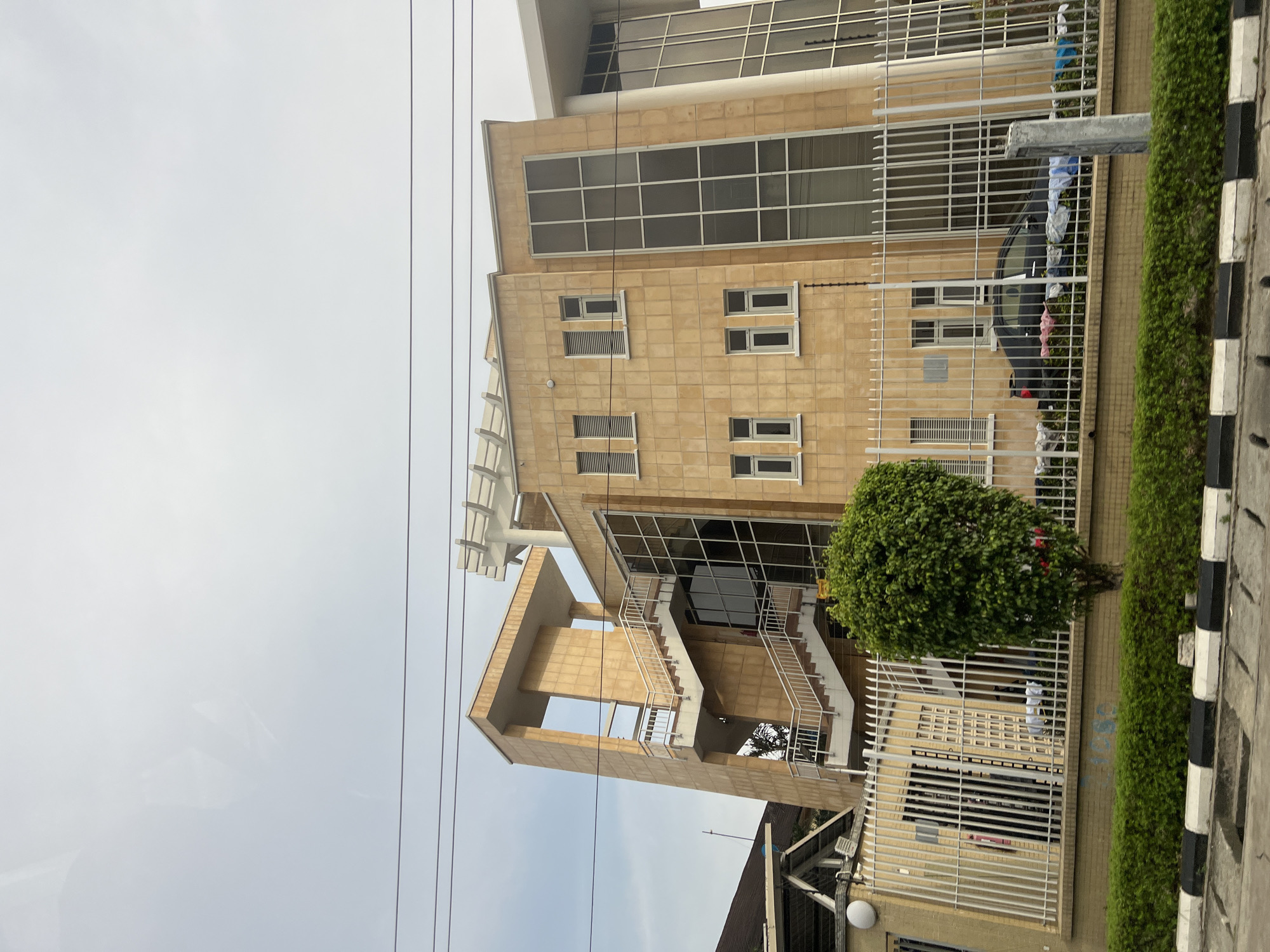 The image depicts a multi-story building in Lekki, Nigeria. The building is constructed with a tan, tiled facade and features extensive glazing. External staircases with white railings are visible, leading to upper floors. A white metal fence surrounds the property, with a strip of green hedge and a small, round bush bordering the sidewalk. A black car is parked behind the fence, and laundry appears to be hanging on the fence. Overhead electrical wires are visible against the overcast sky. The foreground shows a black and white striped curb.