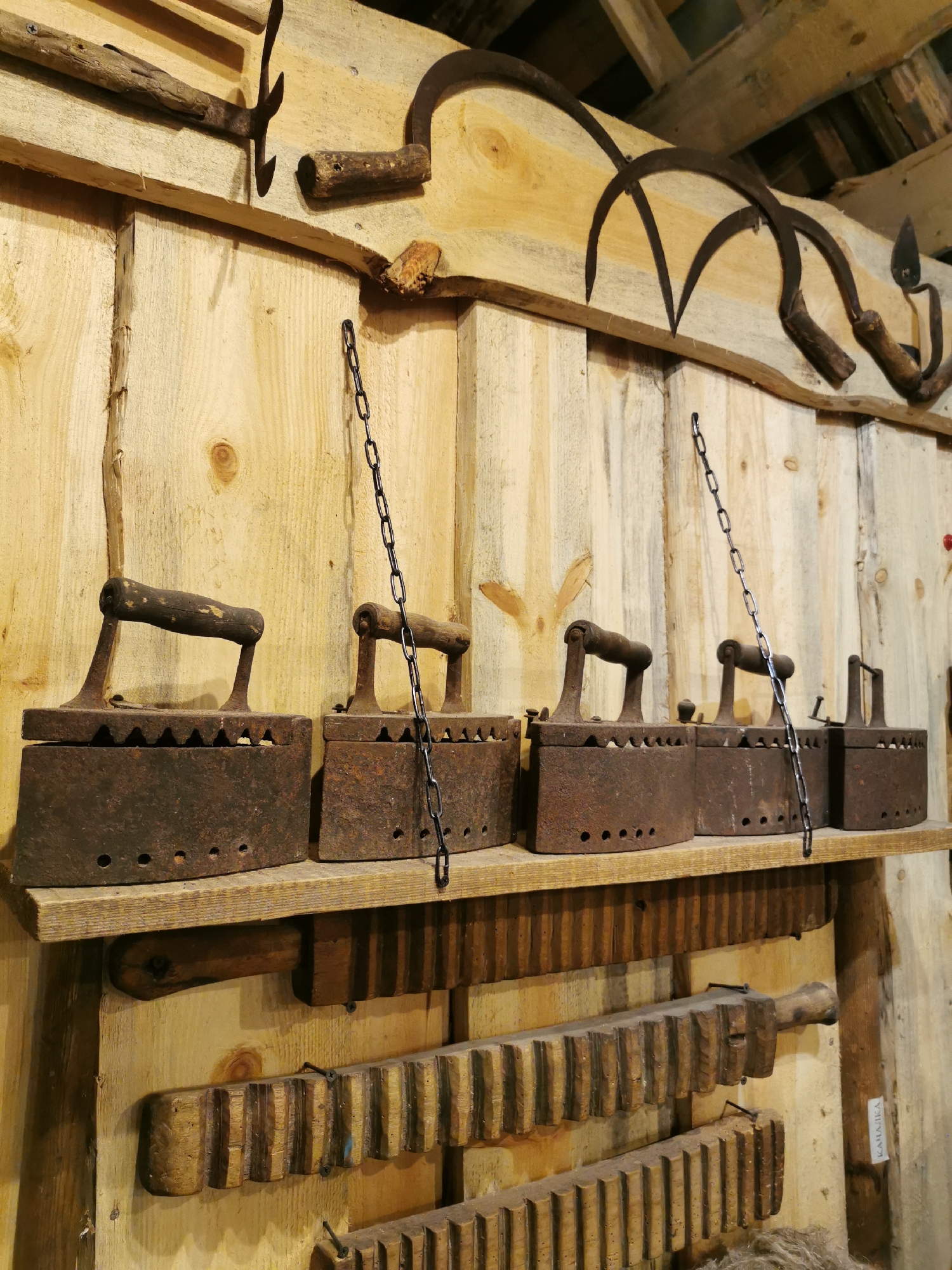 The image shows a collection of vintage agricultural tools and domestic implements displayed on a wooden wall, likely in a museum or historical exhibition space in Шчарчоўскі сельскі Савет, Belarus.
In the upper portion of the image, several old sickles with wooden handles are mounted on the wall. To the right of the sickles, there appears to be a metal farming tool with prongs.
Below these tools, a wooden shelf holds a row of four antique cast-iron clothes irons. Each iron has a wooden handle and a decorative serrated edge. These irons are suspended by metal chains.
Beneath the shelf, two long, wooden implements with parallel, carved grooves are mounted vertically on the wall. These appear to be historical tools, possibly related to processing materials or a form of primitive machinery. The texture and wear on these objects suggest age and frequent use. The overall environment is rustic, with unpainted wooden planks forming the backdrop. A small label with Cyrillic text, "КАHAJJKA," is visible on the right side of the wall. No people are present in the image.