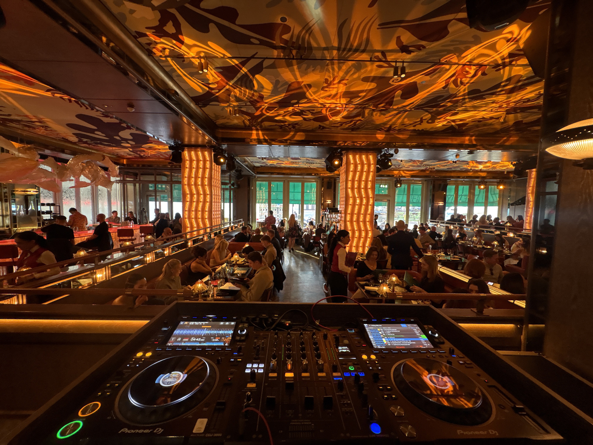 This image shows a restaurant in London, United Kingdom. In the foreground is a Pioneer DJ CDJ-3000 setup. The restaurant has multiple levels with people seated at tables, eating and drinking. The decor includes patterned ceilings and columns with vertical lighting. There are servers attending to the customers. The restaurant is well-lit with both natural light coming through the windows and artificial lighting.