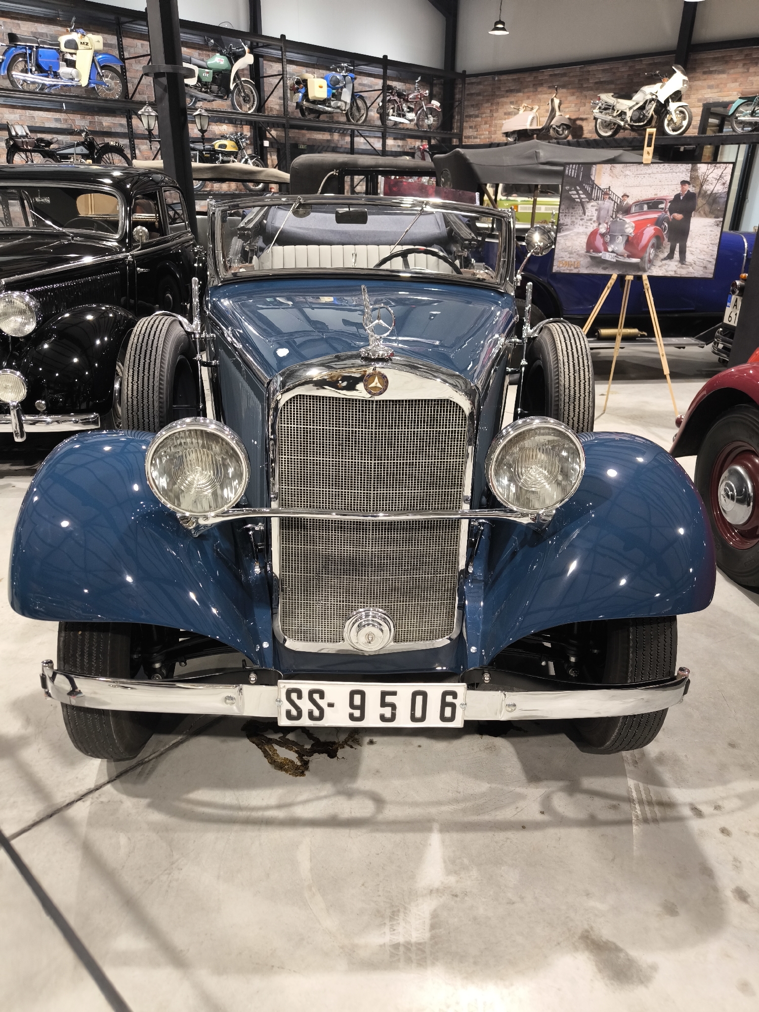 The image shows a blue vintage Mercedes-Benz roadster with the license plate "SS-9506" in what appears to be a museum or showroom. The car is the main subject and is positioned centrally in the foreground. Behind it, there are other vintage cars and motorcycles displayed on shelves. A framed photograph of a red vintage car and two people stands on an easel to the right of the blue car. The floor is a light grey concrete. The location, Kapatovo, Bulgaria, is not visually apparent.