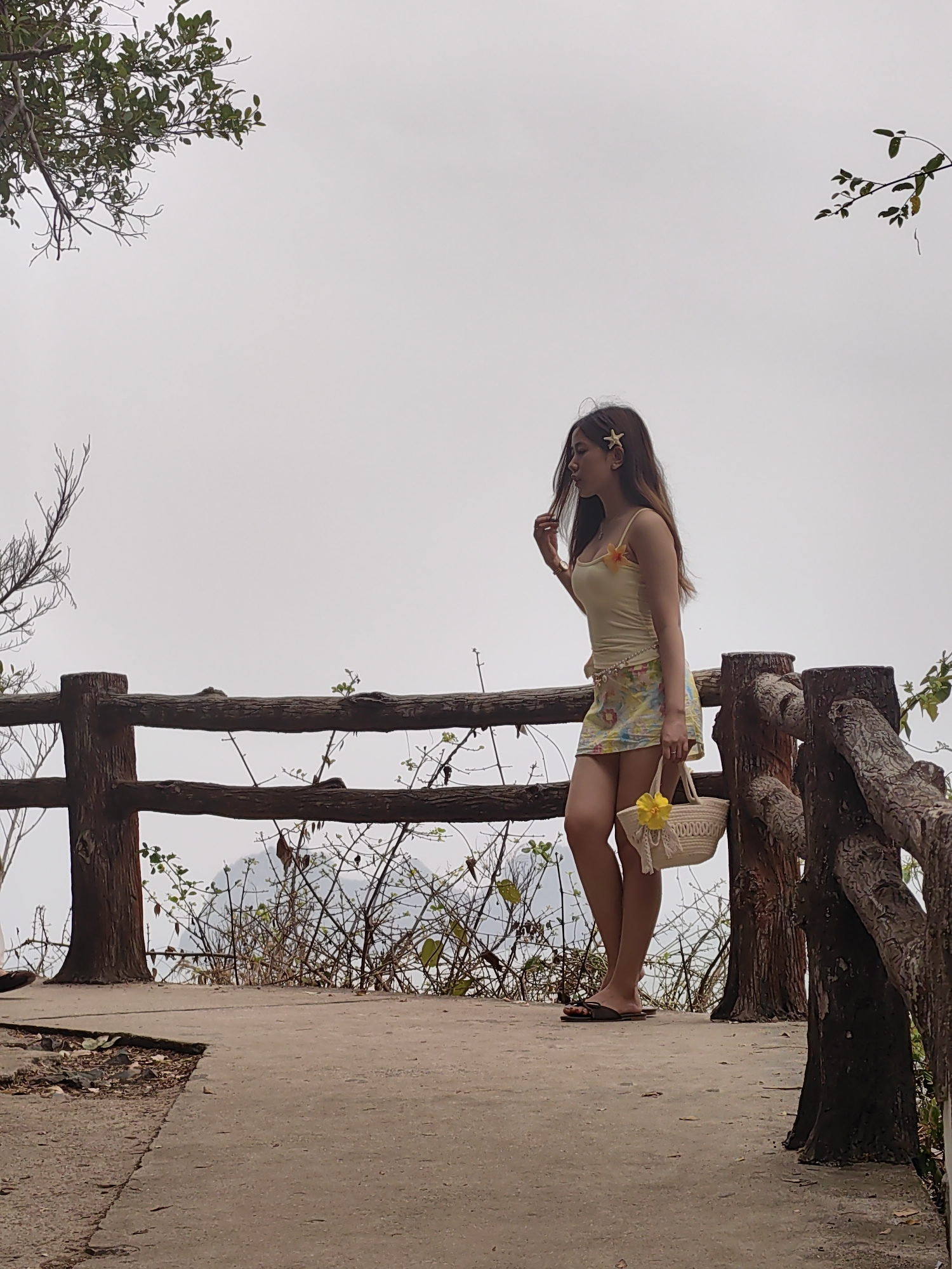 A woman stands on a lookout point in Hải Phòng, Vietnam. The sky is overcast. She is fair-skinned, with long brown hair adorned with a starfish clip. The woman wears a light yellow tank top, a floral skirt, and flip-flops, and carries a small straw bag decorated with a yellow flower. She is touching her hair with her left hand. The lookout has a low, wooden railing with shrubs and trees growing beyond. A concrete path leads to the lookout point.