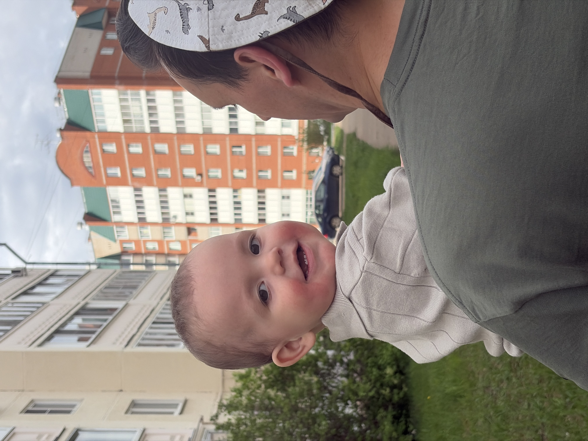 A person is holding a baby in front of an apartment building, likely in Chaikovsky, Russia. The person is wearing a grey-green shirt and a patterned hat. The baby is facing the camera, smiling, and wearing a light-colored, striped shirt. The background shows multi-story apartment buildings in red and white brick with green roofs. There are some trees and a car visible. The sky is overcast.
