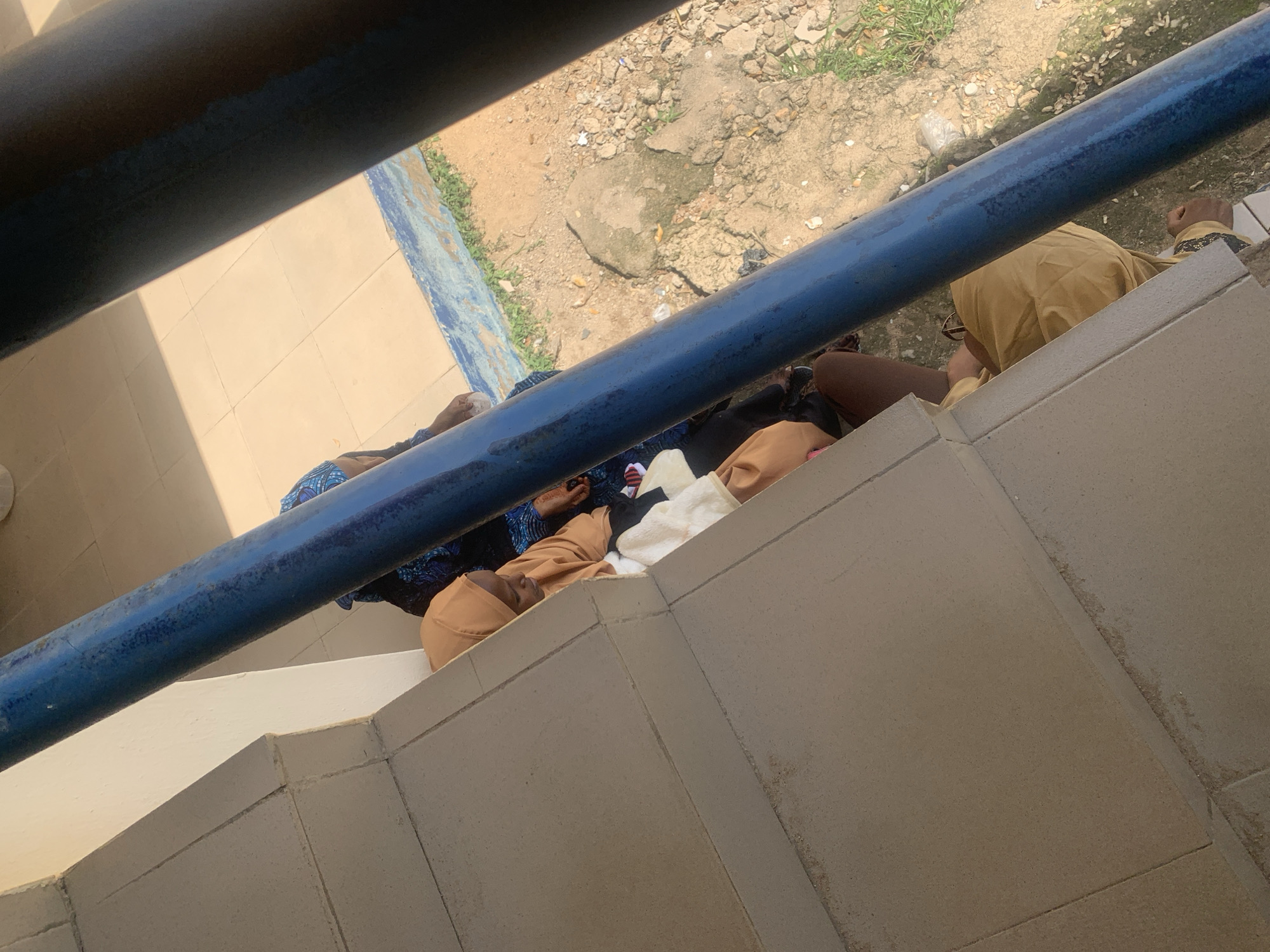 In Jalingo, Nigeria, a group of people are seen resting outdoors on a paved area with a railing. The subjects, dressed in casual attire, appear to be lying down or sitting, some with their eyes closed, suggesting they are sleeping or relaxing. One individual is wearing a tan-colored headscarf and clothing, while another is seen in a blue and black patterned garment. A third person is partially visible in tan clothing.
The scene is set on what appears to be a staircase or raised platform, with beige tiles covering the surface. A prominent blue railing runs horizontally across the frame, partially obscuring the view of the people and the background. Beyond the railing, a dirt and gravel area with sparse green vegetation can be seen, indicating an outdoor environment. The presence of sunlight casting shadows suggests it is daytime. The overall mood is one of relaxation and respite from the heat, possibly during a break. No visible text is present in the image.