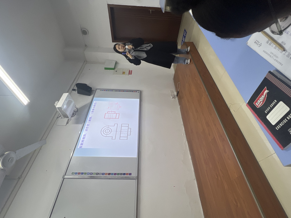 The image depicts an indoor classroom or lecture hall setting, located in Jiangjin, China. A female individual stands on a raised wooden stage at the front of the room, positioned slightly to the right of a large white interactive whiteboard. She wears a black long coat, light blue jeans, and dark sneakers, with a grey scarf draped around her neck, and holds a dark, rectangular object, possibly a remote or pointer, in her hands.
The whiteboard displays a projected image consisting of technical drawings, specifically orthographic projections of a mechanical component, with accompanying Chinese text at the top that translates to "According to two views, draw the third view. (Shape analysis)". A projector is mounted on the ceiling above the whiteboard, alongside wall-mounted speakers. The ceiling also features a white fan and fluorescent light fixtures. The walls are white, showing some discoloration or water staining near the floor level of the stage. A brown door is visible on the far right wall, next to a green panel and red labeled signs. In the lower right foreground, the back of a student's head is partially visible. On a blue student desk in the foreground, an open "EUROPA STIFF COVER EXERCISE BOOK" and another open textbook with a pencil are present.