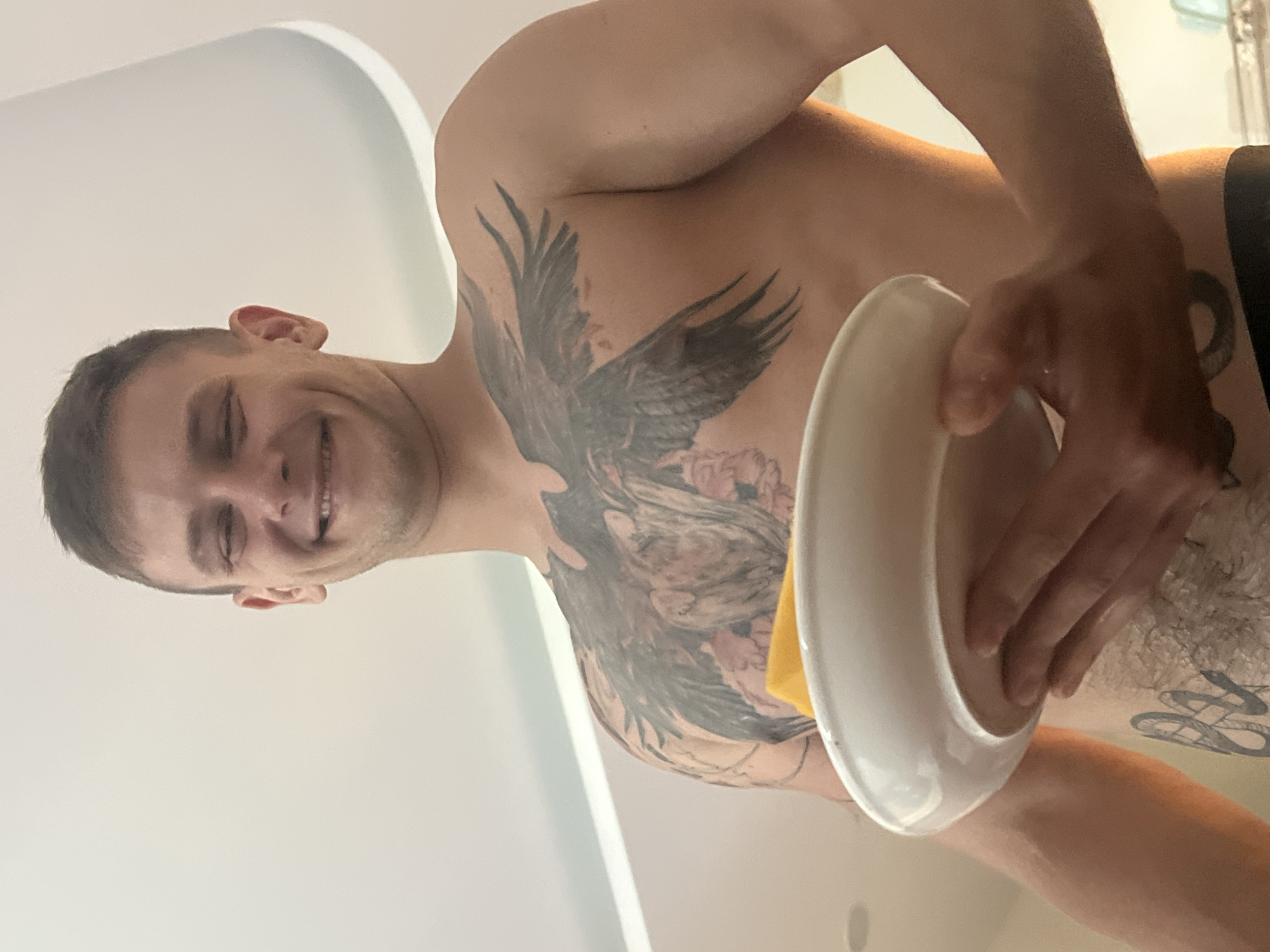 A shirtless man with tattoos is holding a white plate with a yellow object inside. The background is a white wall and ceiling. The location is not discernable from the image.