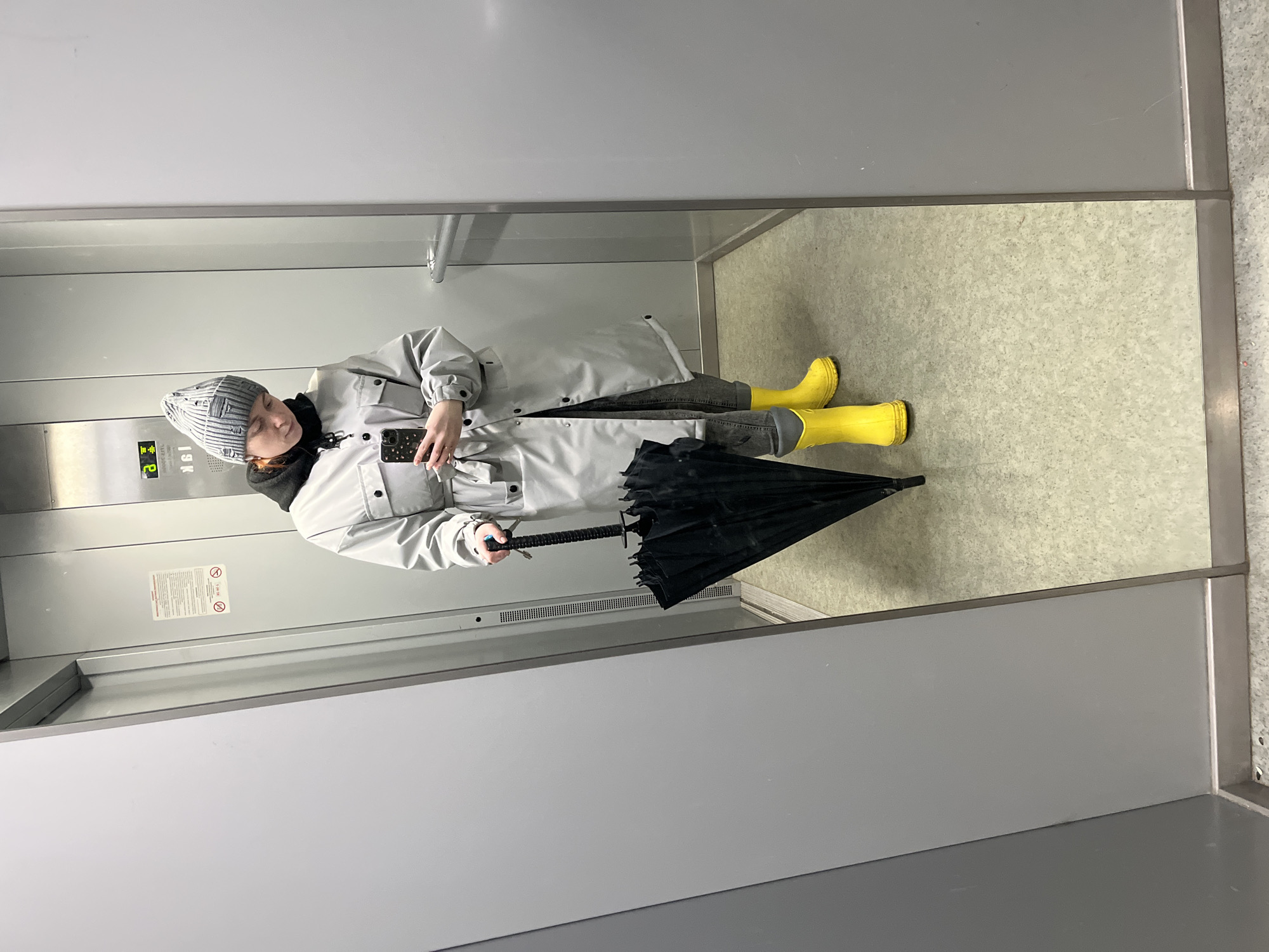 A person stands inside an elevator, wearing a gray coat, gray beanie, and yellow rain boots. They hold a black umbrella and a phone. The elevator has metal walls and a light-colored floor. Elevator buttons are visible on the left wall. The location is inside an elevator, unknown if it is in Chaikovsky, Russia.