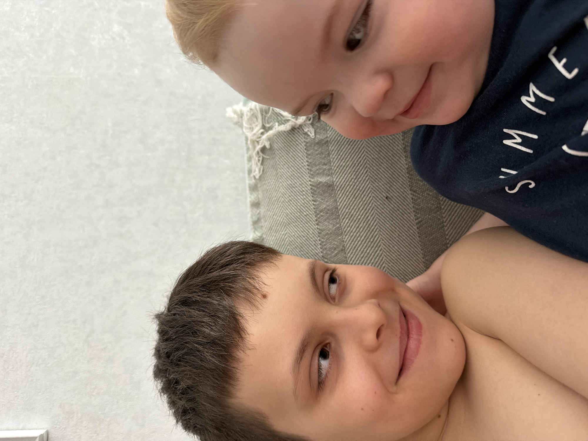Two young children are depicted in a close-up, intimate overhead shot, lying down indoors. The child at the top of the frame has light blonde hair and fair skin, wearing a dark blue t-shirt with the word "SUMMER" printed in white capital letters. Their gaze is directed downwards with a soft, affectionate expression. The second child, positioned lower in the frame, has dark brown hair and fair skin, and appears shirtless, revealing their shoulder. This child looks upwards with a warm, broad smile, returning the gaze of the first child, suggesting a strong bond between them.
Between the children is a textured fabric, possibly a cushion or blanket, with a green or greyish-green herringbone pattern. The remaining background is a light-colored, subtly textured surface, likely a wall or sheet. The bright, even lighting indicates an indoor daytime setting. The overall mood of the image is tender and joyful, capturing a moment of connection between the two. The provided location context is Votkinsk, Russia.
