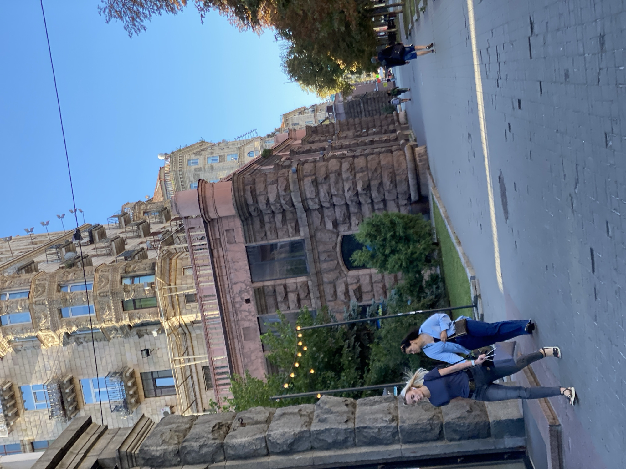 In the city of Unknown, Unknown, on a clear, sunny day, two women walk along a pedestrian path lined with trees and buildings. The woman in the foreground, with blonde hair, wears a dark blue t-shirt and dark grey jeans, carrying a light grey shopping bag. Beside her, a woman with dark hair wears a light blue button-down shirt and dark blue pants, carrying a small brown purse. A man in a dark t-shirt and blue shorts walks away from the camera further down the path, while other pedestrians are visible in the distance. The architecture features a large, stone-faced building with ornate balconies and windows, and a lighter-colored, multi-story building behind it. The sidewalk is paved with grey stones, and a string of lights hangs between poles on the left side of the path.