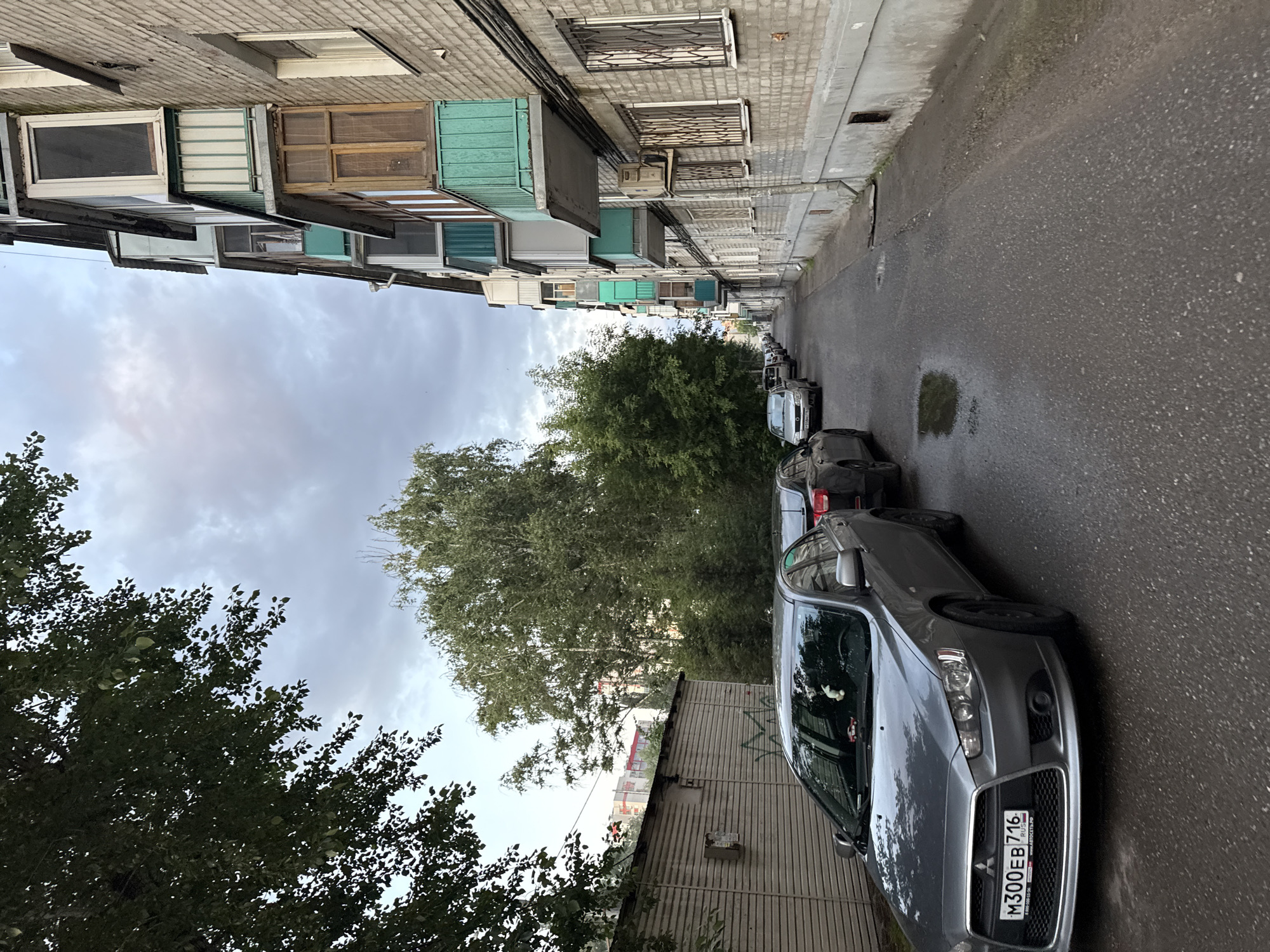 An exterior, medium-shot, eye-level photograph captures a street scene in Kazan, Russia. The image is dominated by a gray four-door sedan parked on the left side of a narrow asphalt street. The sedan's license plate is partially visible, reading "M300EB 716." Further down the street, additional vehicles are parked in a single line on both sides, receding into the distance. To the right of the street, a multi-story brick apartment building stands tall, featuring several balconies with green and wooden accents. The building's facade is weathered, and metal grates cover some of the windows. To the left of the street, a long, light-colored corrugated metal building with a graffiti tag is visible. Dense green trees line the background, with more apartment buildings peeking through the foliage. The sky above is overcast with gray clouds. The asphalt street appears damp, with a small puddle reflecting the sky. The overall scene depicts a typical urban residential area.