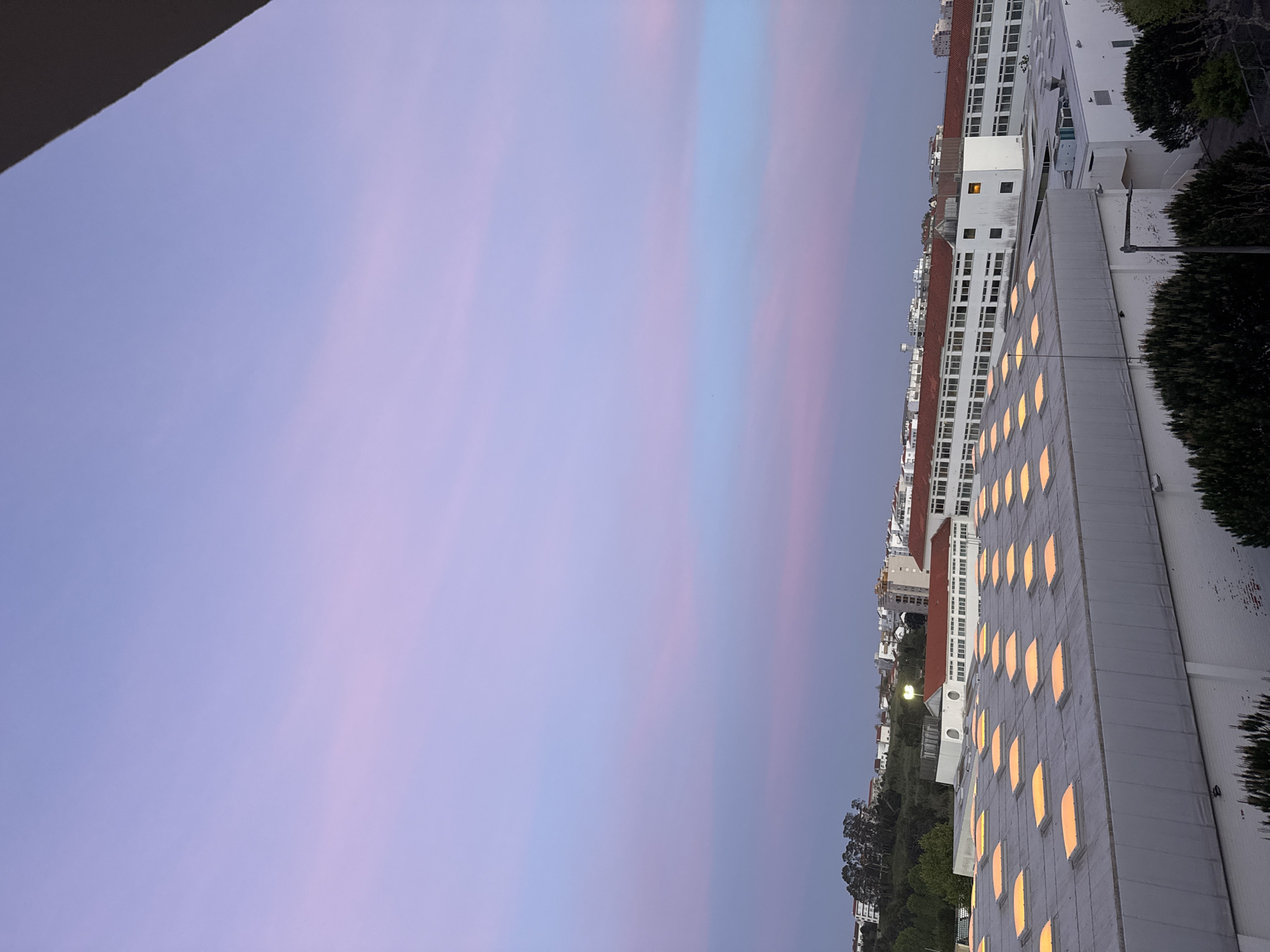 The image captures an urban landscape in Figueira da Foz, Portugal, during twilight, likely at sunset. The sky, which occupies a significant portion of the frame, is a soft blend of lavender and periwinkle blue, interspersed with faint, elongated streaks of pinkish-orange and light blue clouds, reflecting the fading or emerging light of the day.
On the right side of the scene, several buildings are visible. In the mid-ground, a large, multi-story structure with a light grey or white facade dominates, featuring numerous rectangular windows uniformly arranged. These windows emit a warm, golden-yellow glow, indicating that interior lights are turned on. Behind this building, other white buildings with distinctive red-tiled roofs can be seen stretching into the distance, some also showing illuminated windows. Dark green trees and bushes are scattered throughout the scene, particularly in the lower right corner and nestled among the buildings.
A dark, angled architectural element, possibly an overhang or part of an adjacent building, partially frames the upper left corner of the sky. The overall atmosphere is tranquil and serene, with the artificial lights of the buildings complementing the soft, natural colors of the twilight sky. No people or specific activities are visible, and there is no discernible text within the image. The weather appears clear with minimal cloud cover.