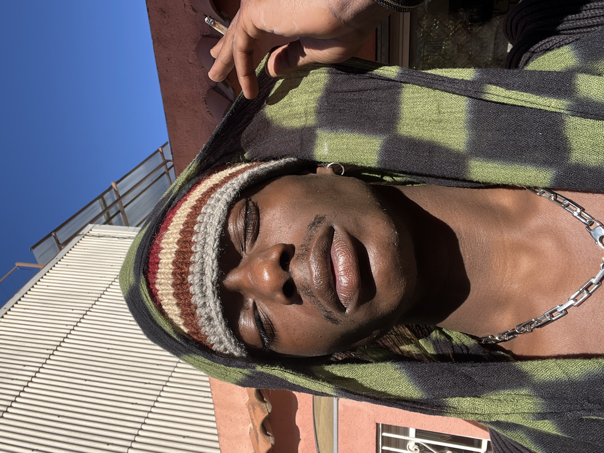 A close-up, low-angle shot depicts a Black person with eyes closed, wearing a striped knit cap and a hooded garment with a green and black tie-dye pattern. The person has a silver chain necklace and a small hoop earring. Above the person's hand holds a cigarette, with a portion of a building visible in the upper left. The environment includes a building facade and what appears to be the sky. The location cannot be determined from the image.