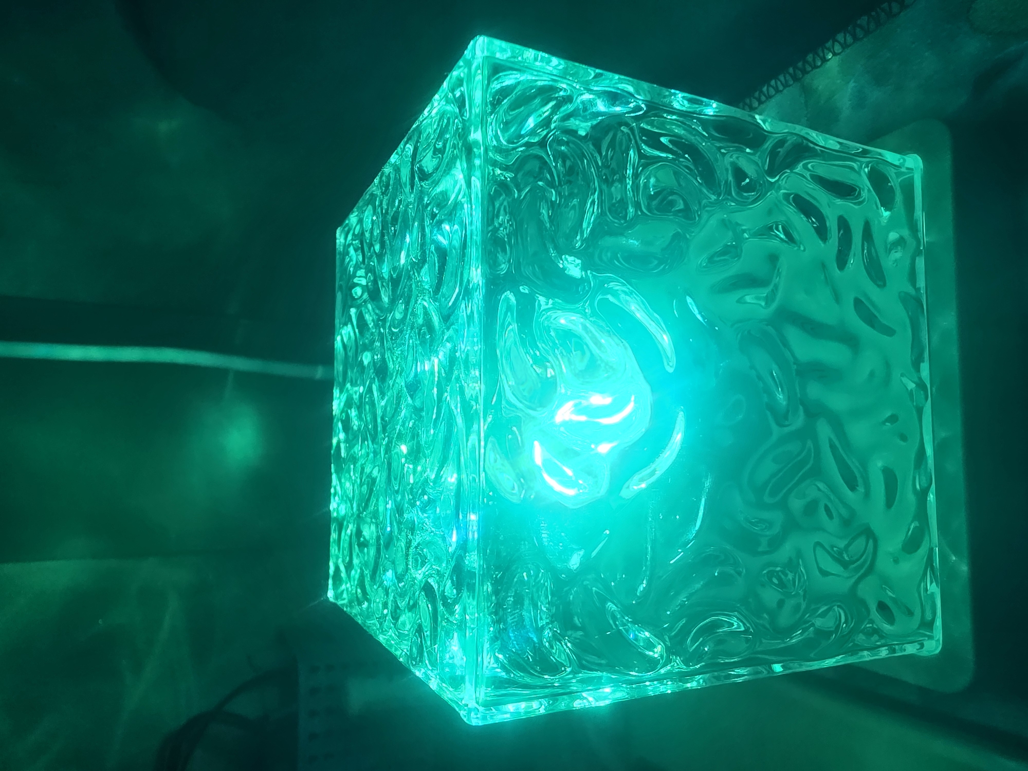 A close-up, low-angle shot captures a cube-shaped lamp emitting a vibrant green light. The lamp's surface has a textured, organic pattern that refracts the internal glow, creating a dynamic visual effect. The background is dark and out of focus, with faint green light reflecting on a wall, suggesting the lamp is the primary light source in a dimly lit room. The scene is devoid of human presence or any specific context related to Bila Tserkva, Ukraine.
