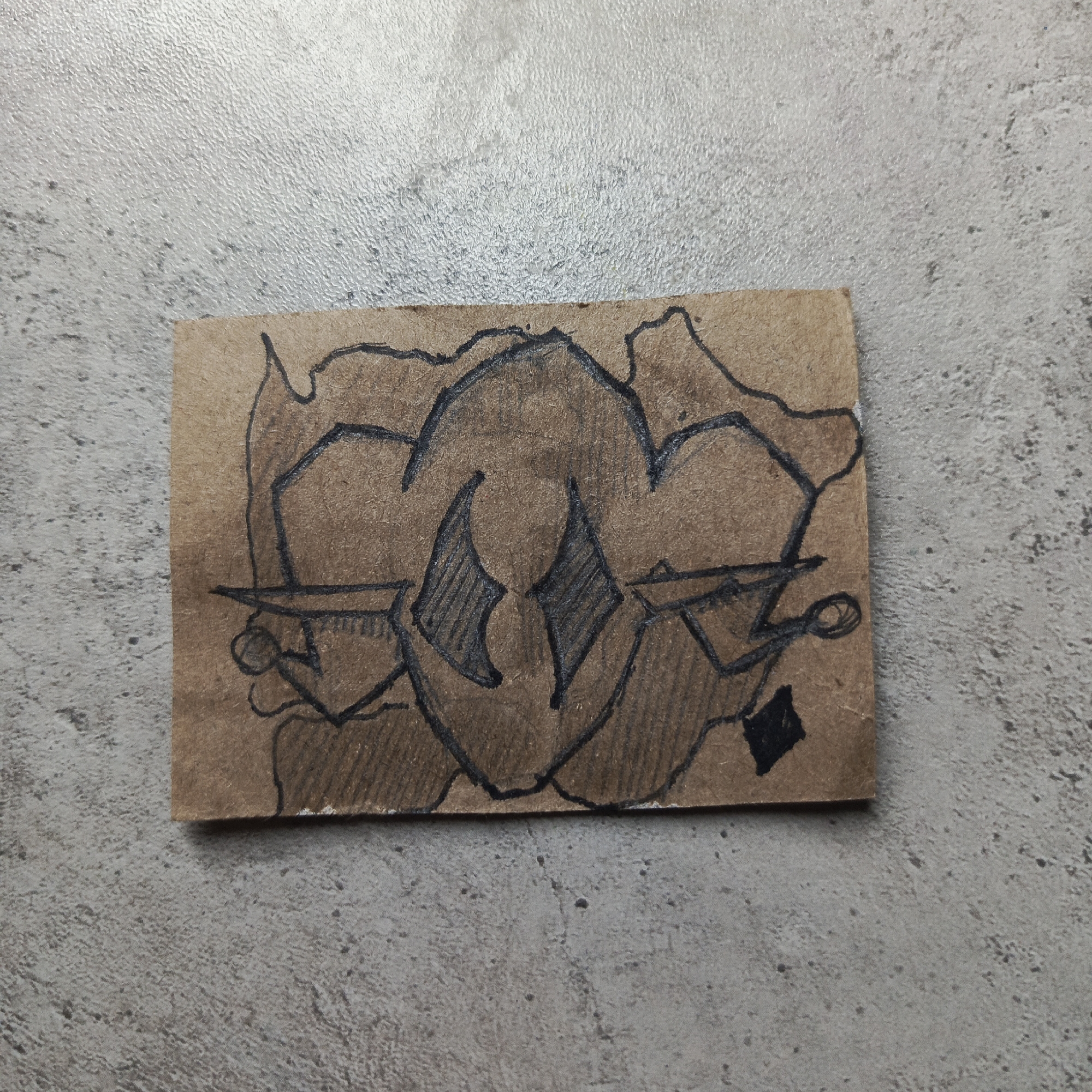 A small, rectangular piece of brown paper with a black ink drawing sits on a gray, textured surface. The drawing appears to be an abstract, symmetrical design. The central figure includes two shaded diamond shapes. Outlines include curved lines and some geometric shapes. There is a solid black square in the bottom right corner. The location of the paper is Unknown, Unknown.