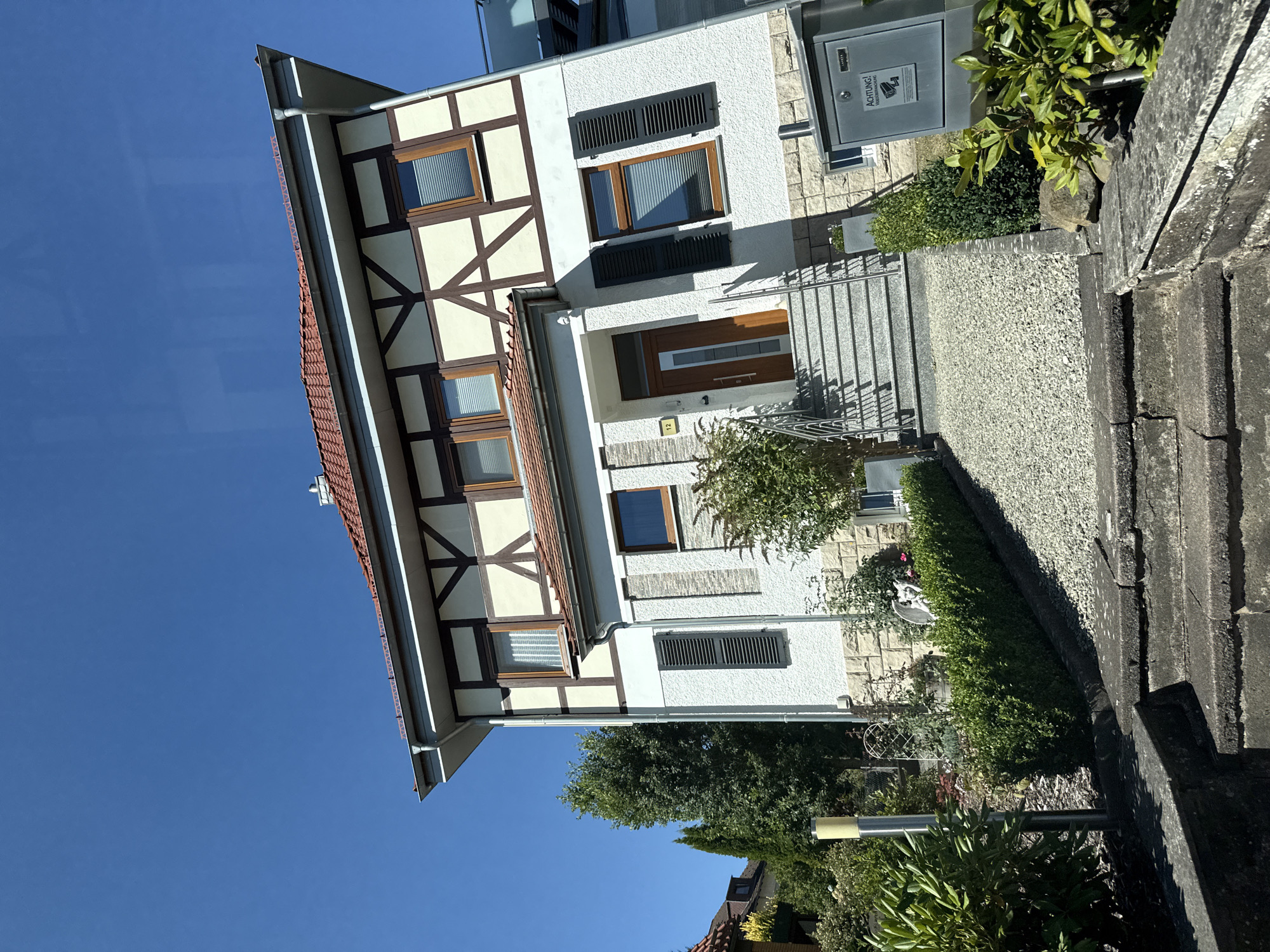 A full shot of a multi-story home in Bad Sachsa, Germany. The home has a traditional half-timbered upper story with white infill and dark wood beams, topped by a red tile roof. The lower story is white stucco over a stone base. A set of stone stairs leads up to the front door, which is made of wood with a glass panel. Shutters are closed on the windows. A mailbox is mounted on the side of the house. The yard is landscaped with bushes and trees. The sky is clear and blue.