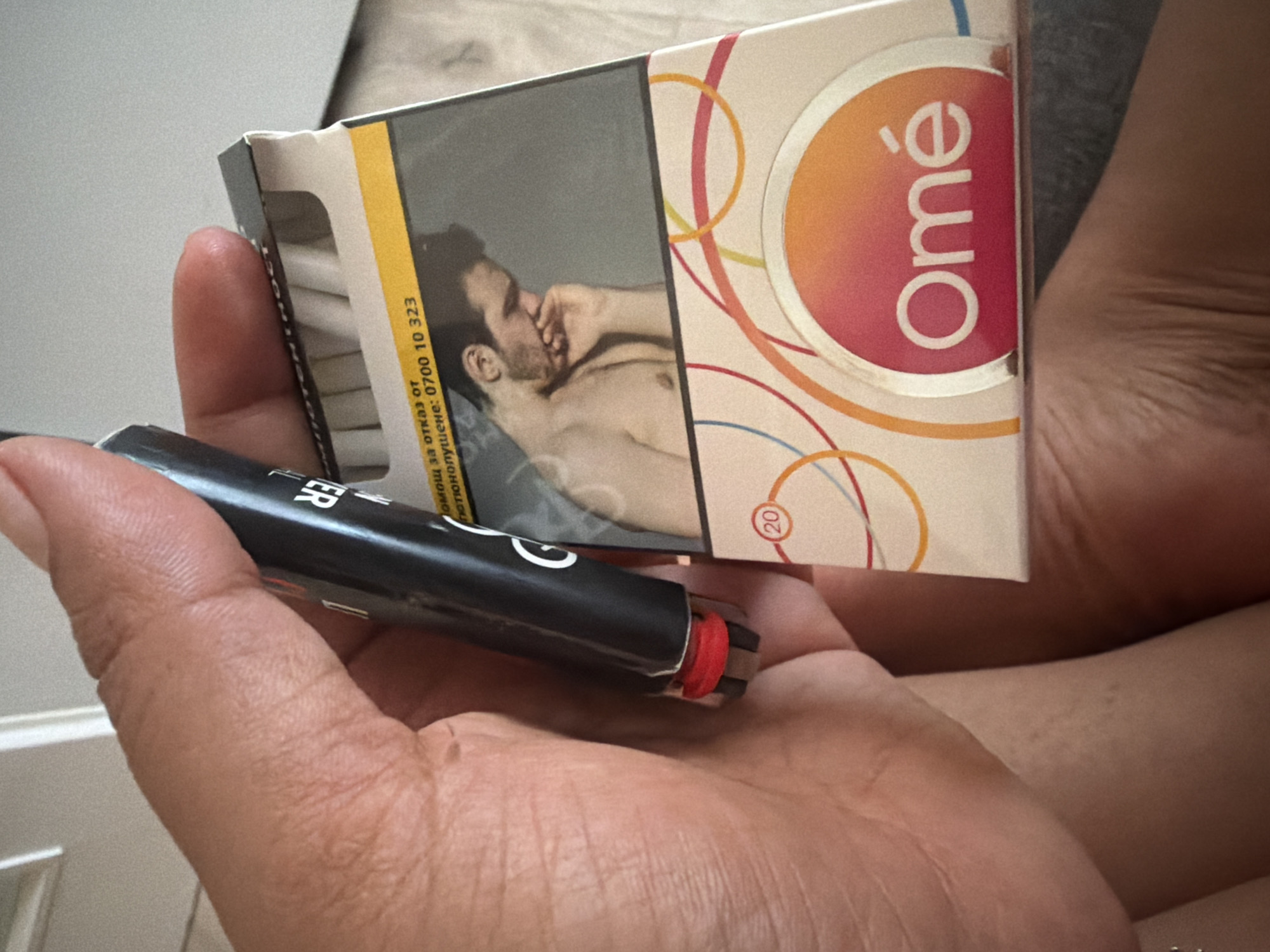 A person's hand holds a black lighter with a red tip and a pack of cigarettes. The cigarette pack is white with a yellow band across the top. It features an image of a shirtless man and the brand name "Omé" in a circular logo with a gradient of pink and orange. Text on the pack reads "Помощ за отказ от" and "Телефонна линия: 0700 10 323". The cigarettes inside the pack are visible. The background appears to be an indoor setting with a light-colored wall and a wooden floor. The image does not provide any information about the location within the city of Duisburg, Germany.