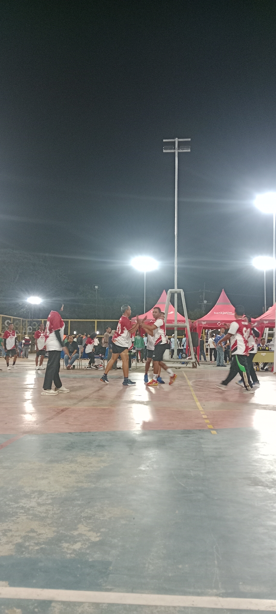 The image depicts an outdoor volleyball match at night in Tuban, Indonesia. A group of athletes, primarily wearing red and white uniforms, are actively engaged in the game on a court illuminated by floodlights. In the background, spectators are visible, some seated and others standing. Red tents with the "bankjatim" logo are set up behind the court, suggesting sponsorship for the event. A tall light pole with multiple lights stands prominently in the center of the frame, casting shadows on the court. The court itself appears to be a hard surface, possibly concrete or asphalt, with painted lines marking the boundaries. The overall atmosphere suggests a community sporting event taking place in the city of Tuban.