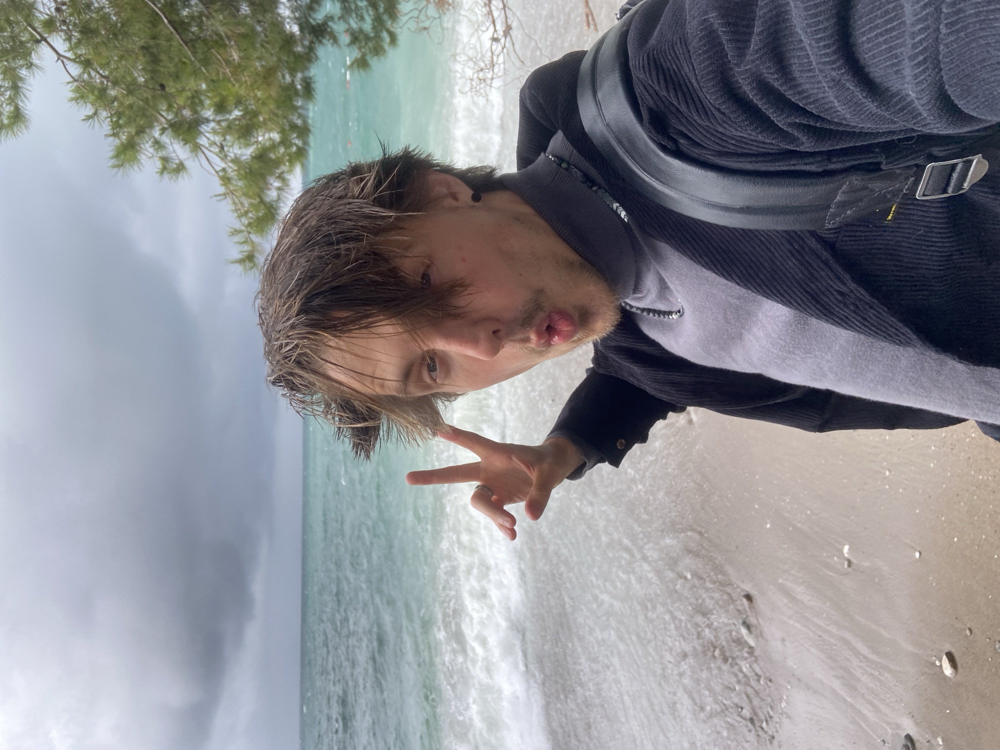 A selfie of a young man on a beach in Bar, Montenegro. He is making a peace sign with his right hand and has his lips pursed. He wears a gray sweater underneath a dark, corduroy jacket and has a black bag strap over his shoulder. The beach is sandy and wet from the water. The sea is aqua in color, with a storm approaching on the left side of the frame. A tree is visible in the upper left corner of the image.