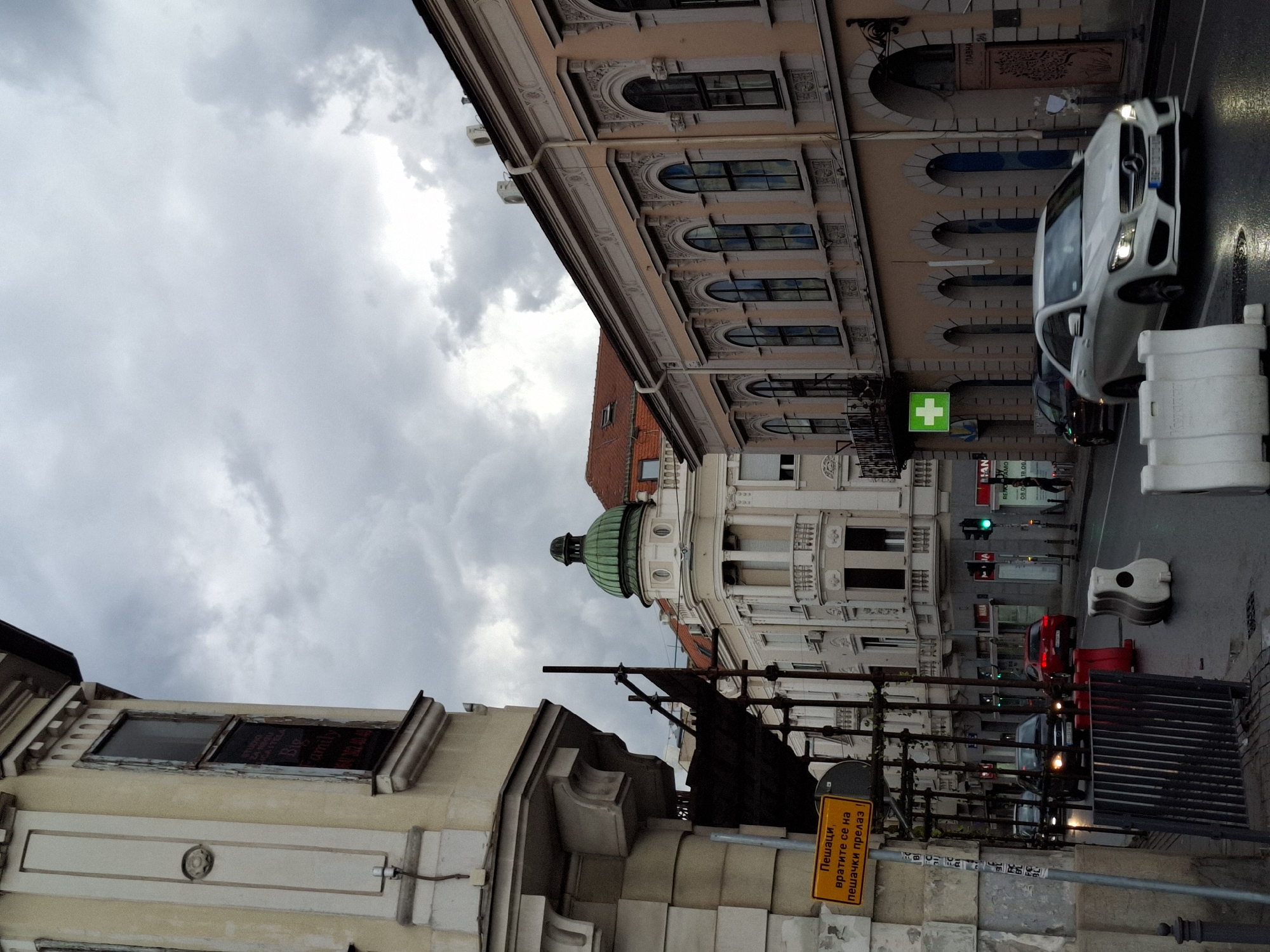 The image captures a street scene in Zemun, Serbia, under a cloudy sky. The foreground features a light-colored Mercedes-Benz driving on a wet asphalt road, with its headlights on. Several white plastic traffic barriers are present on the roadside. To the left, a building with beige-colored facade and intricate architectural details is visible, with scaffolding on its side. A yellow warning sign with black text is affixed to a pole near this building. Further down the street, other buildings with classical architecture line the road. Red and black cars are seen in the middle ground. A green cross sign is mounted on the facade of one of the buildings on the right. The overall impression is of a city street during daylight, possibly after or during rainfall.