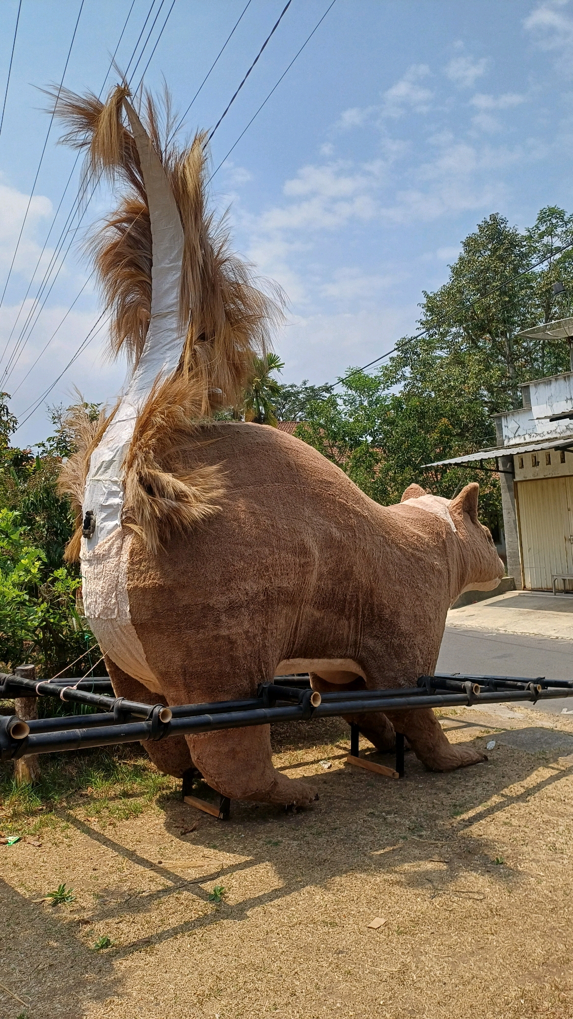 A large, brown, furry sculpture of an animal, possibly a bear or lion, is positioned outdoors on a dirt and grass surface. The sculpture has a long, bushy tail made of straw-like material. It is supported by a black metal frame structure. In the background, there are green trees, power lines, a cloudy blue sky, and a building with a light-colored facade and closed doors. The location is identified as Karangwuni, Indonesia.