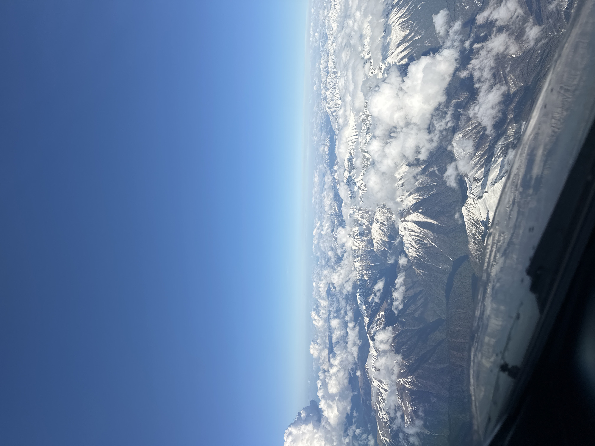 The image shows a view from an aircraft window. On the right, a portion of the aircraft window is visible. Beyond the window, there is a landscape of snow-capped mountains interspersed with clouds. The left side of the image shows a clear blue sky. There is no indication of the image being in Khimki, Russia.