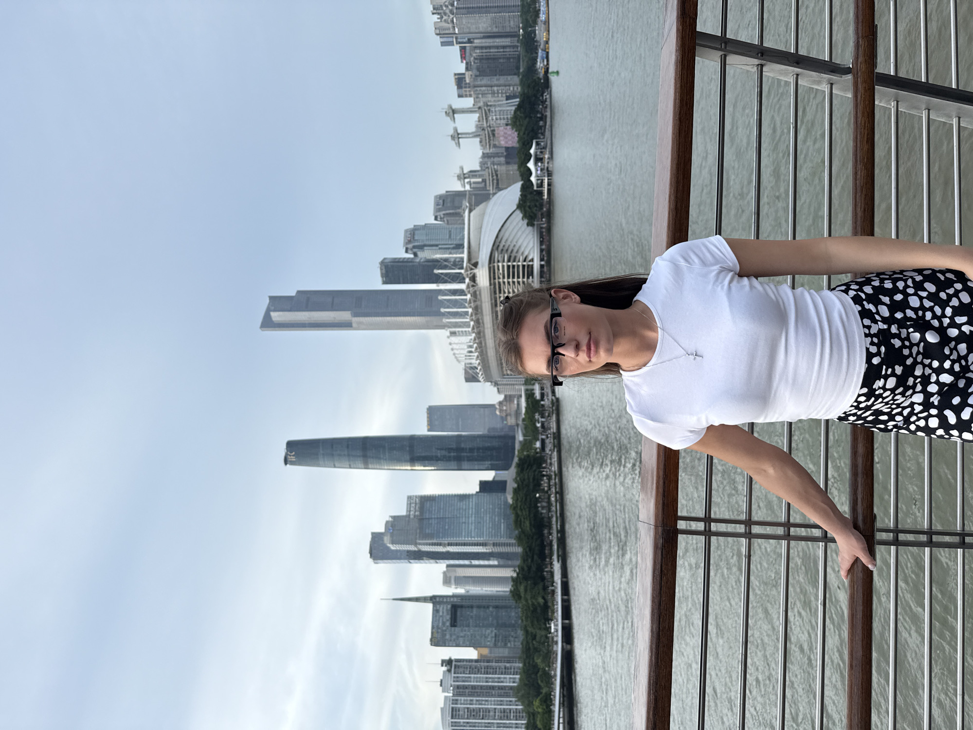 The image depicts a young woman leaning against a railing with a cityscape in the background. She is wearing a white t-shirt and a black skirt with white polka dots, and glasses. Her arm is resting on the railing, and she is looking towards the camera.
The cityscape features numerous skyscrapers of varying heights and architectural styles, indicating an urban environment. A body of water, likely a river or bay, separates the foreground from the city. The sky is overcast, suggesting a cloudy day. The overall scene appears to be a scenic viewpoint, possibly a promenade or observation deck, offering a panoramic view of the city. The woman's posture and gaze suggest a relaxed or contemplative mood. There is no visible text in the image. Based on the context provided, the location is likely Yuexiu District, China.